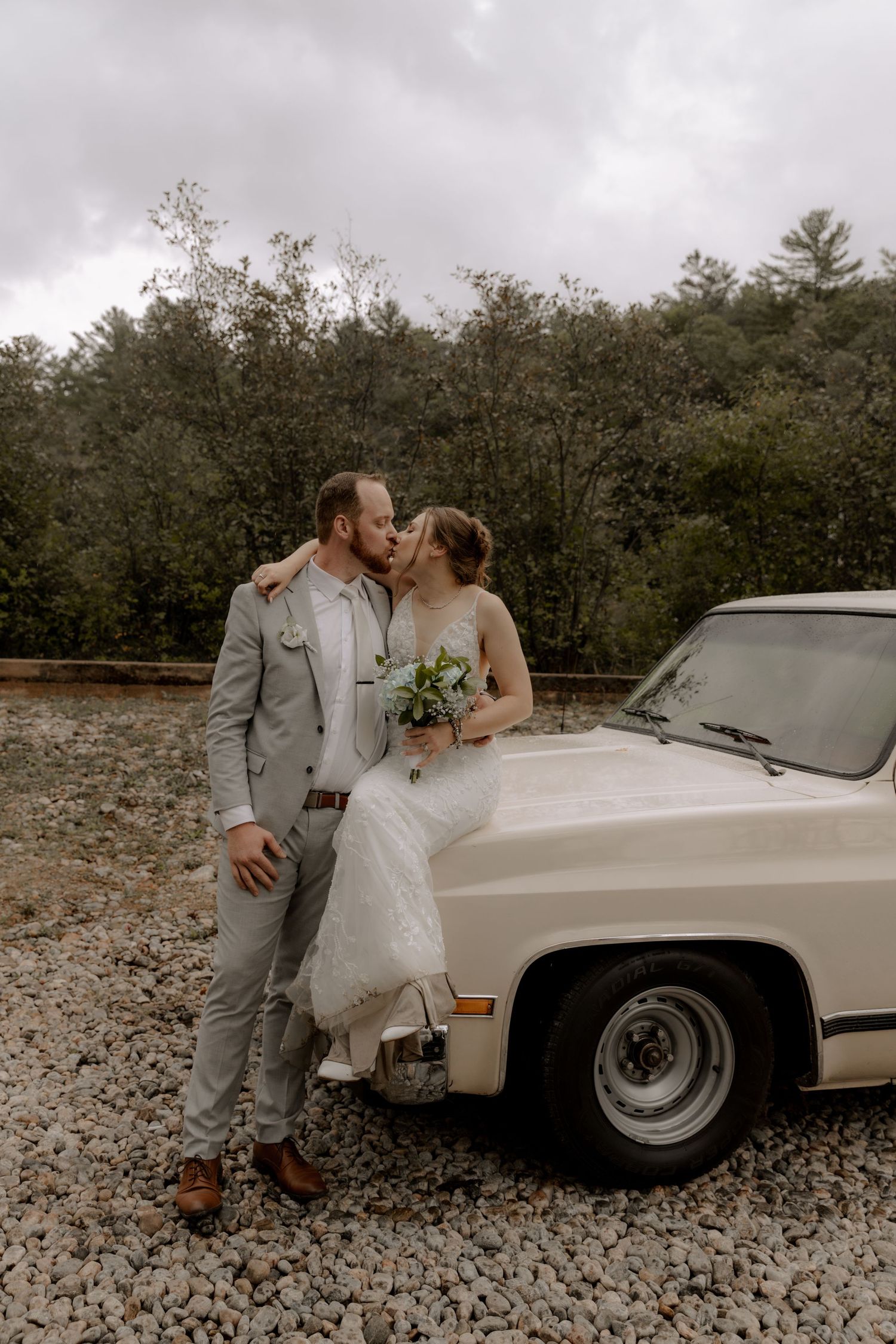 A romantic wedding couple poses next to a vintage car on a gravel road surrounded by trees.