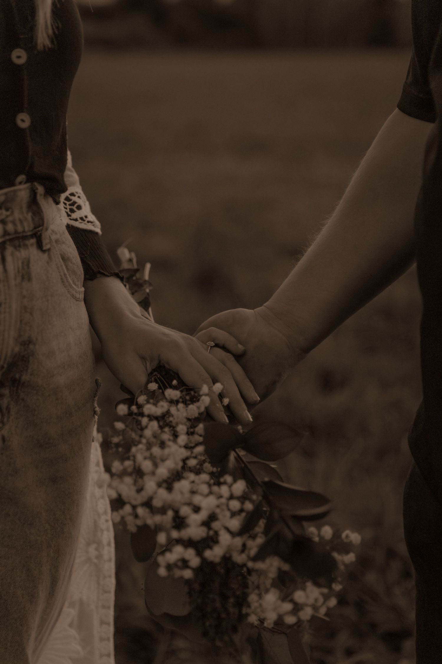 A sepia-toned close-up of two hands clasping together against a blurred floral background.