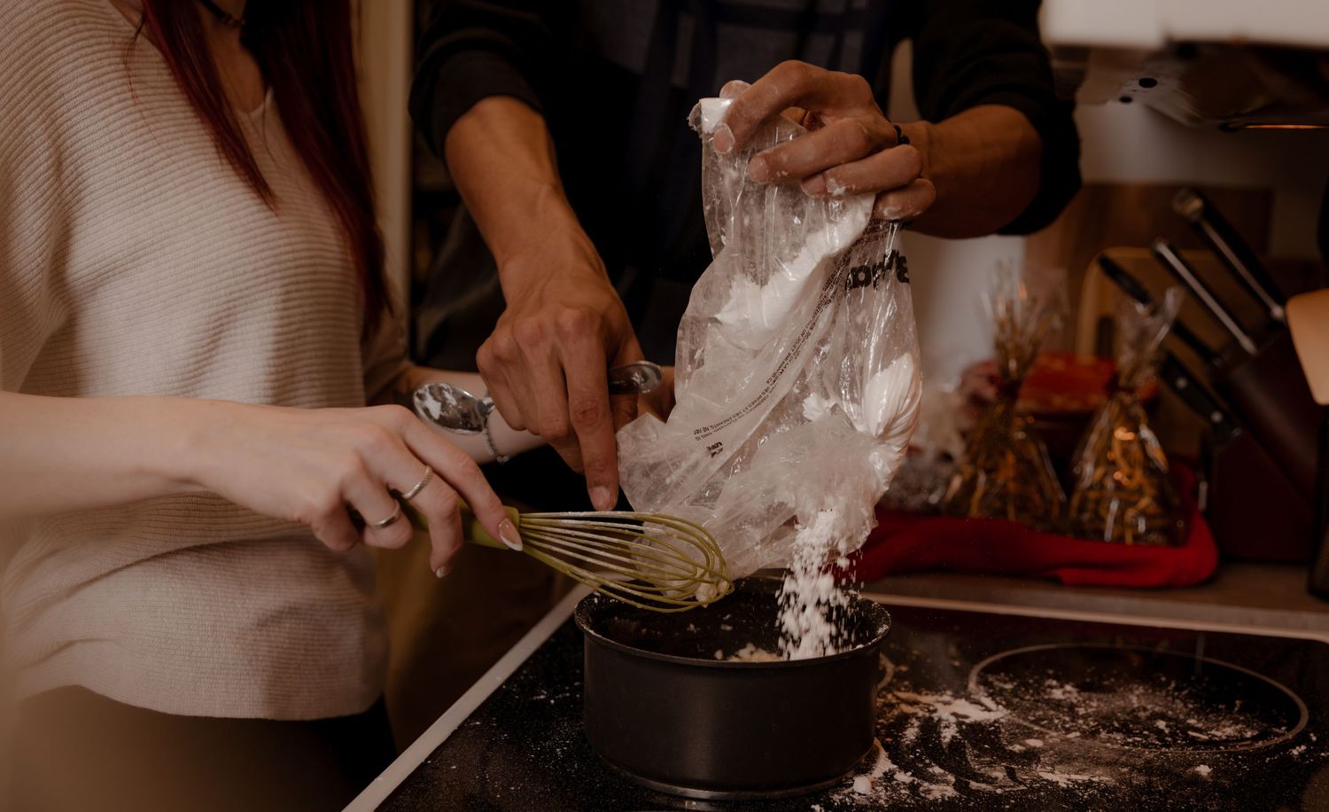 Flour being poured into a cooking pot while hands prepare ingredients in a warm-lit kitchen.