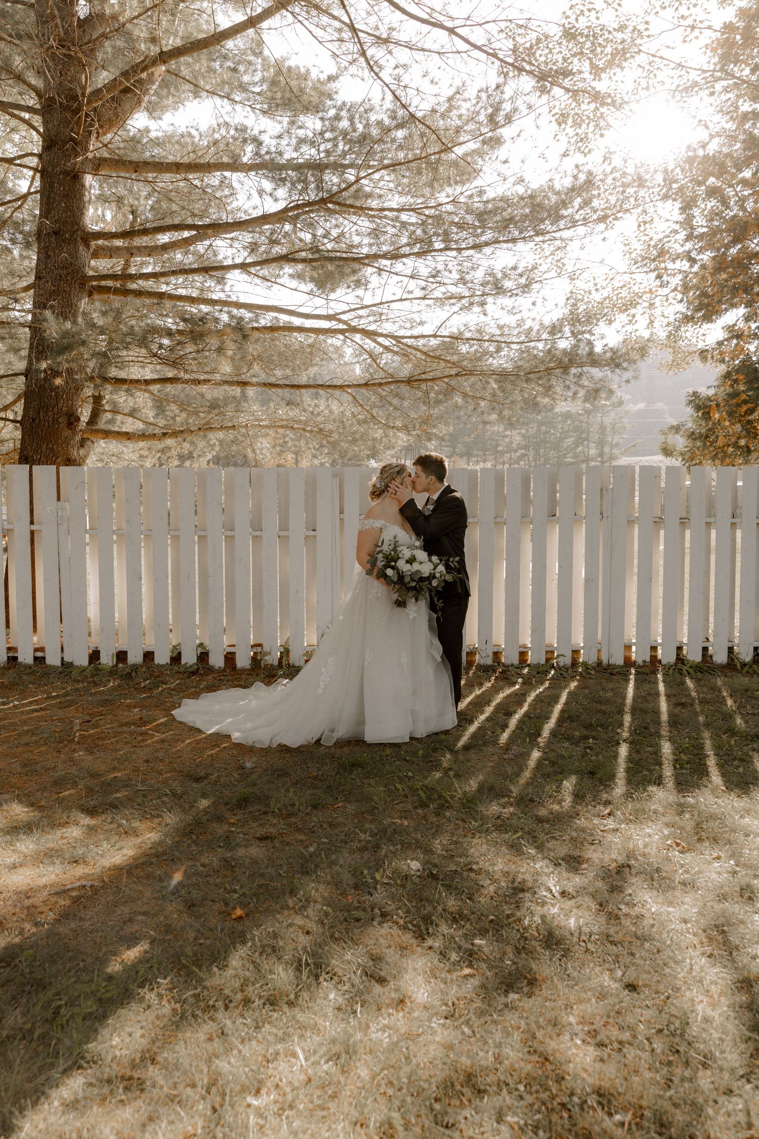 A wedding couple poses against a white picket fence with sunlight filtering through pine trees above.