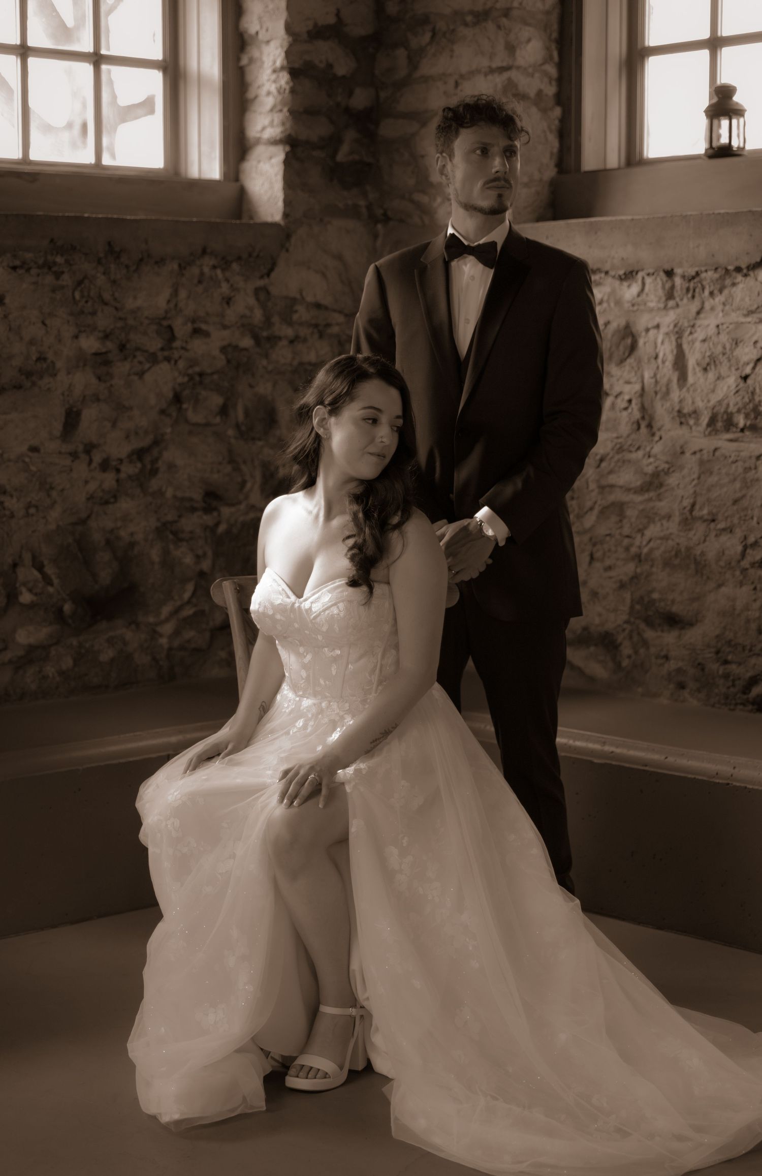 Sepia-toned formal wedding portrait in rustic stone room with window light.