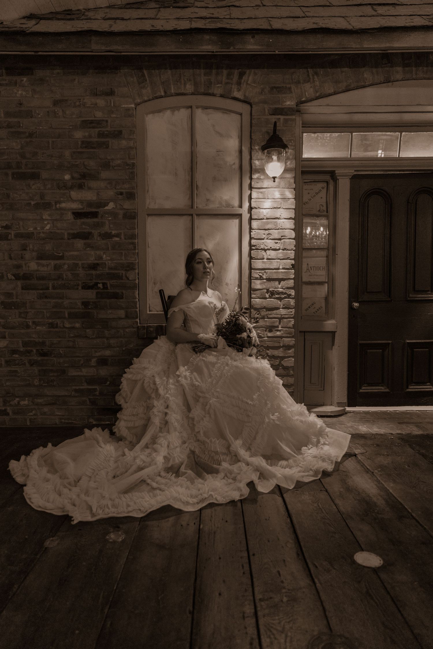 Bride in elaborate wedding gown posed on wooden floor against brick wall with vintage window.