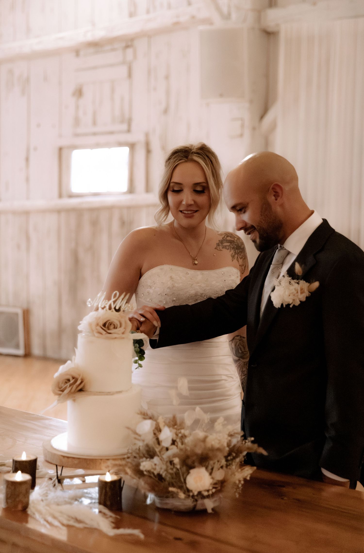 A couple cuts their elegant white wedding cake together in a rustic white barn setting.