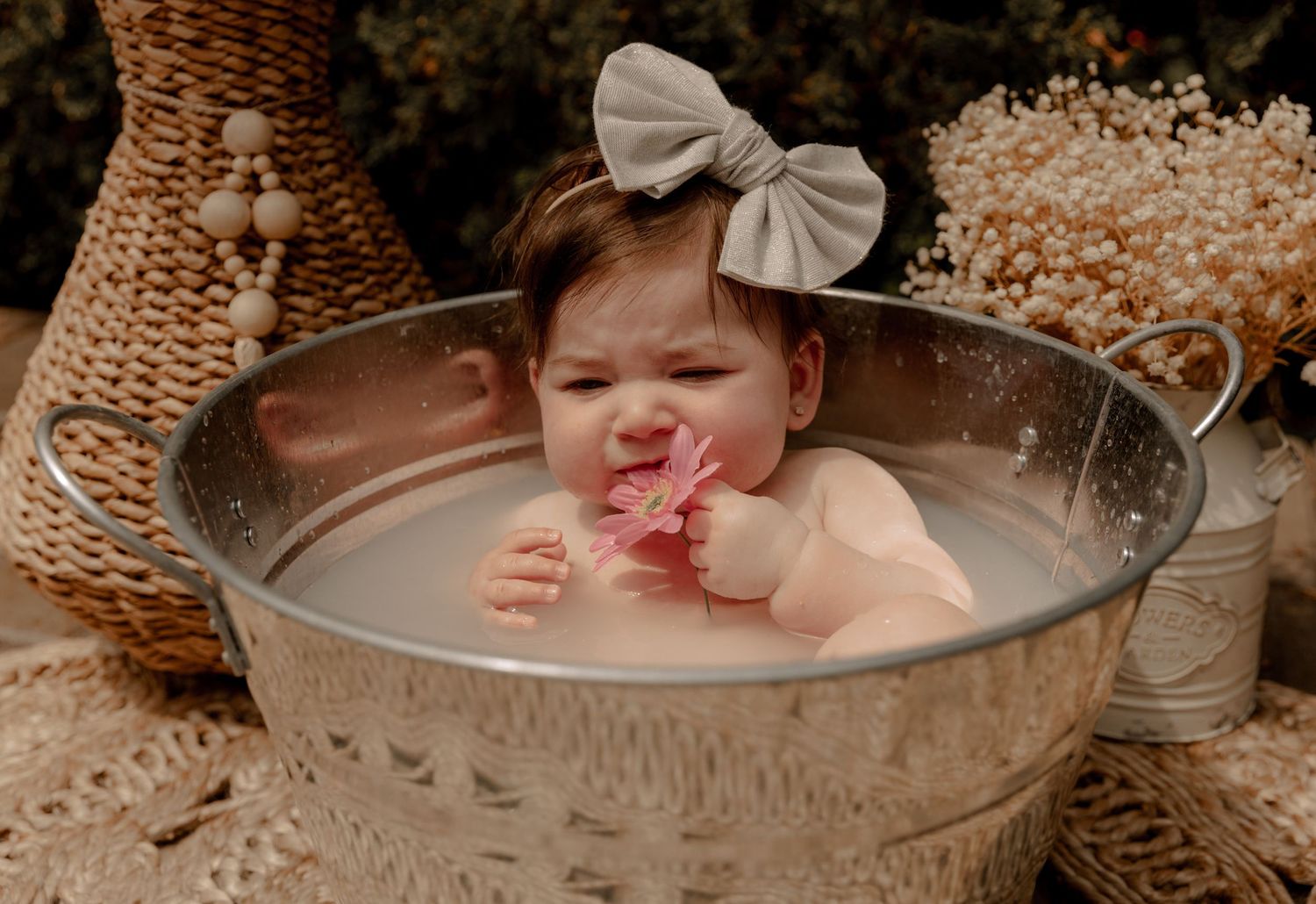 Baby wearing a gray bow sits in a vintage galvanized metal tub during a rustic outdoor photo session.