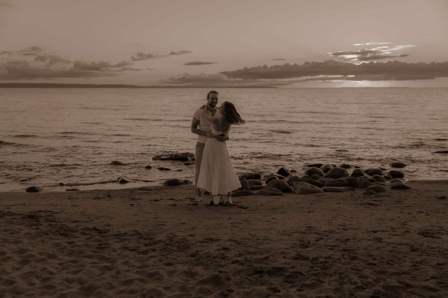 Black and white photo of couple embracing on beach at sunset.