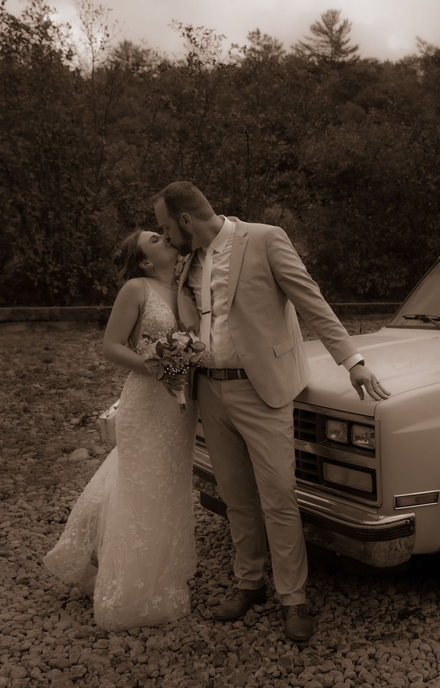A sepia-toned wedding photo of a couple embracing next to a classic car.