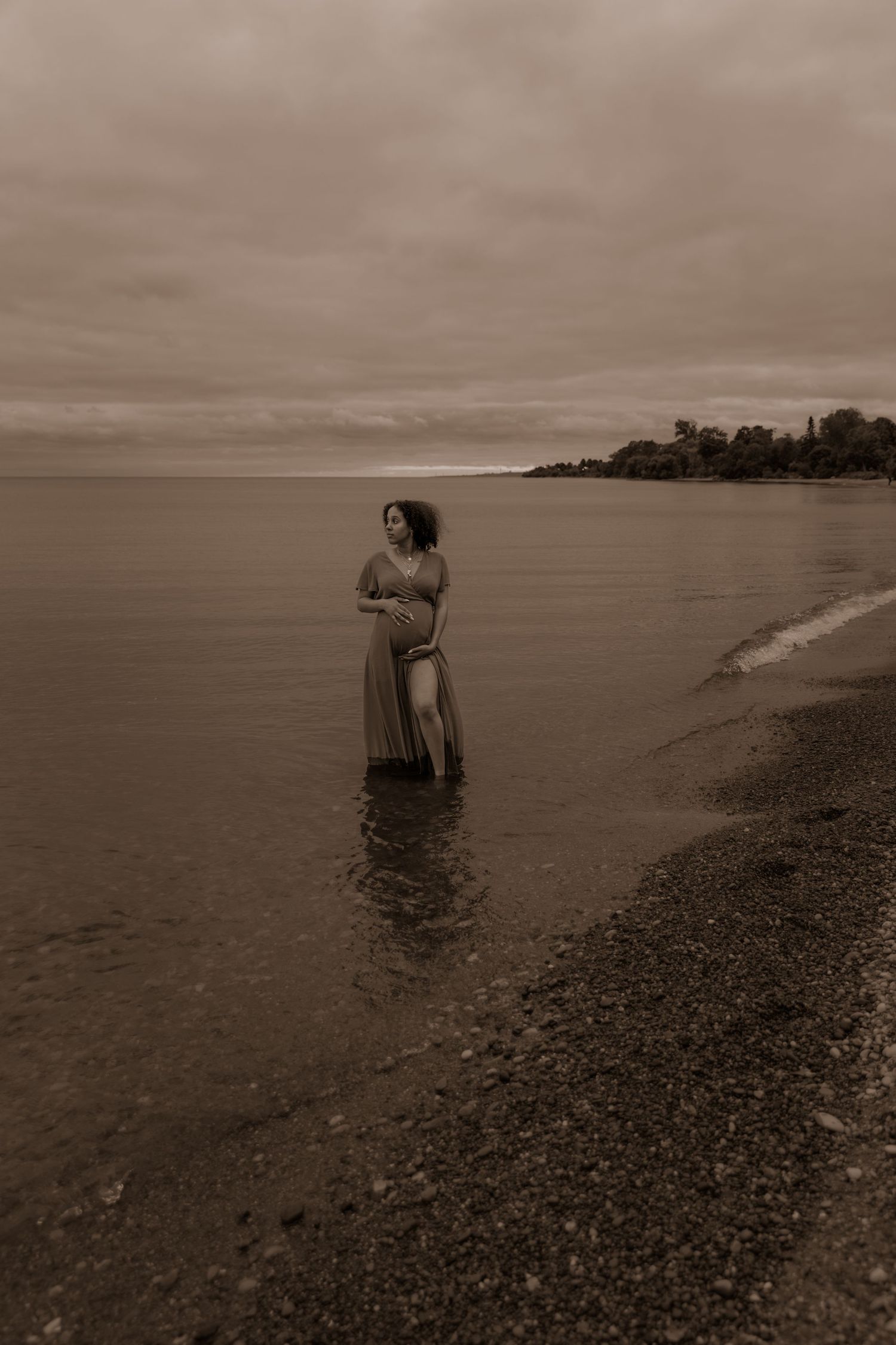 A sepia-toned photograph shows a figure in a flowing dress standing in shallow water along a rocky shoreline.