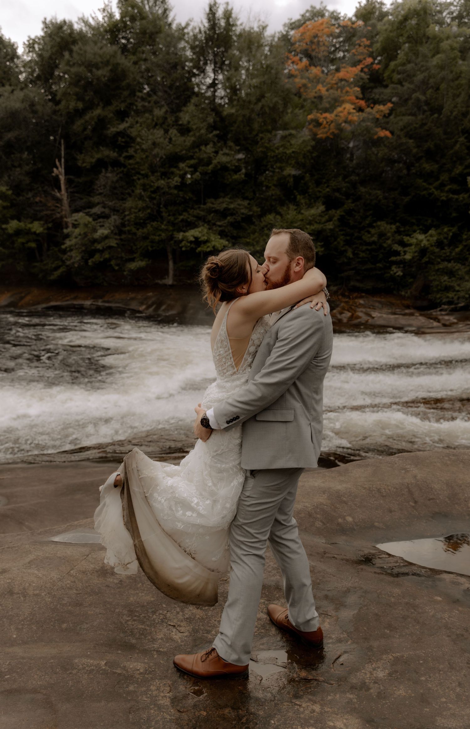 A romantic wedding couple embraces by rushing water and autumn trees wearing gray suit and white dress.