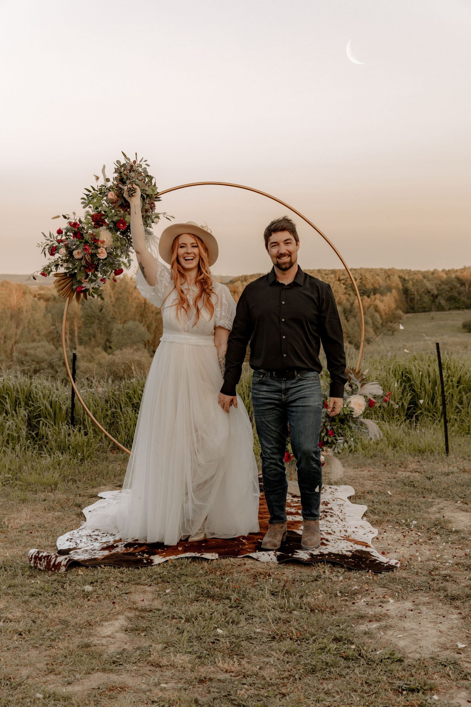 Wedding couple standing under floral arch against sunset sky in rustic outdoor setting.