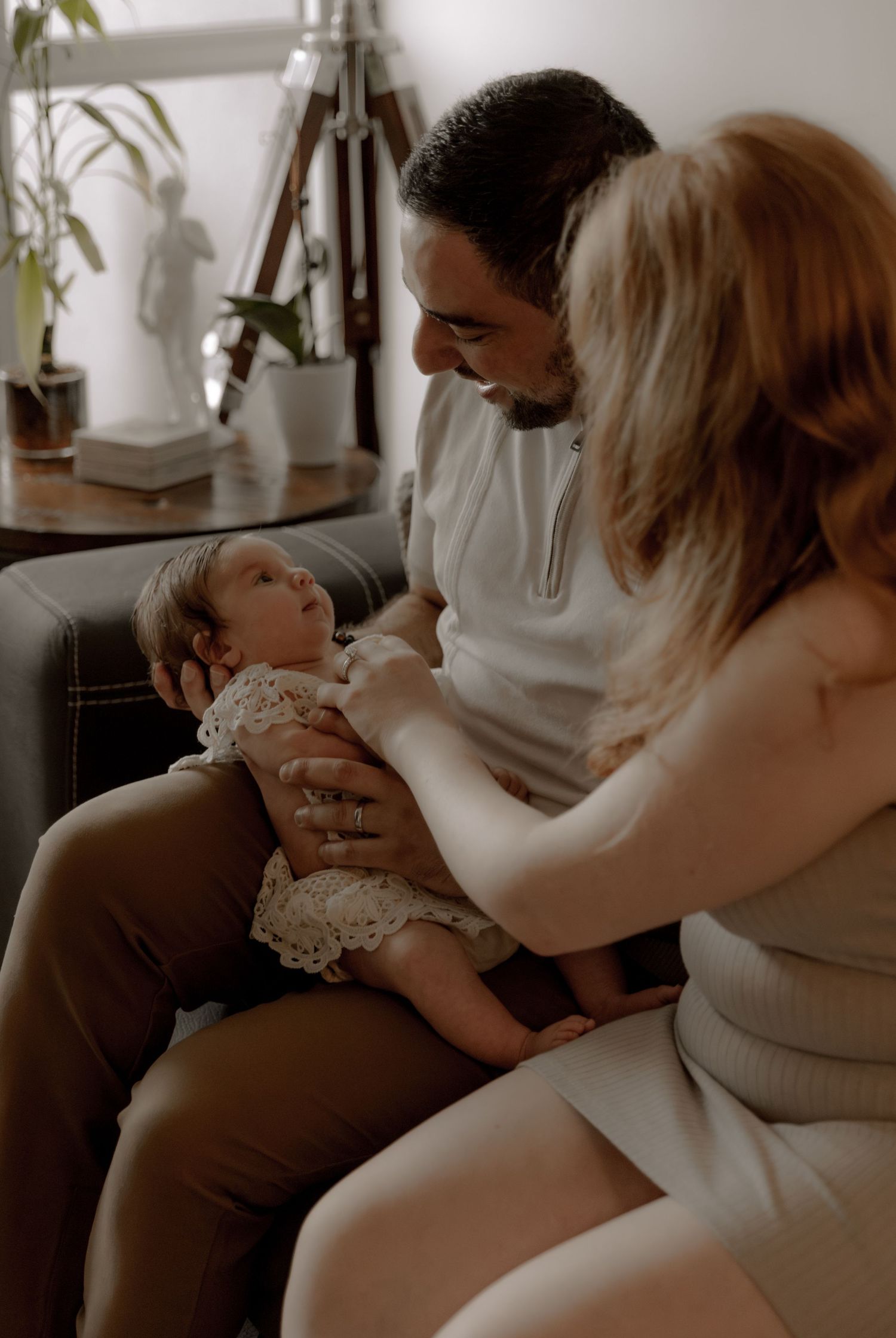 A tender family moment as new parents share a loving interaction with their small baby on a couch in a warm-lit room.