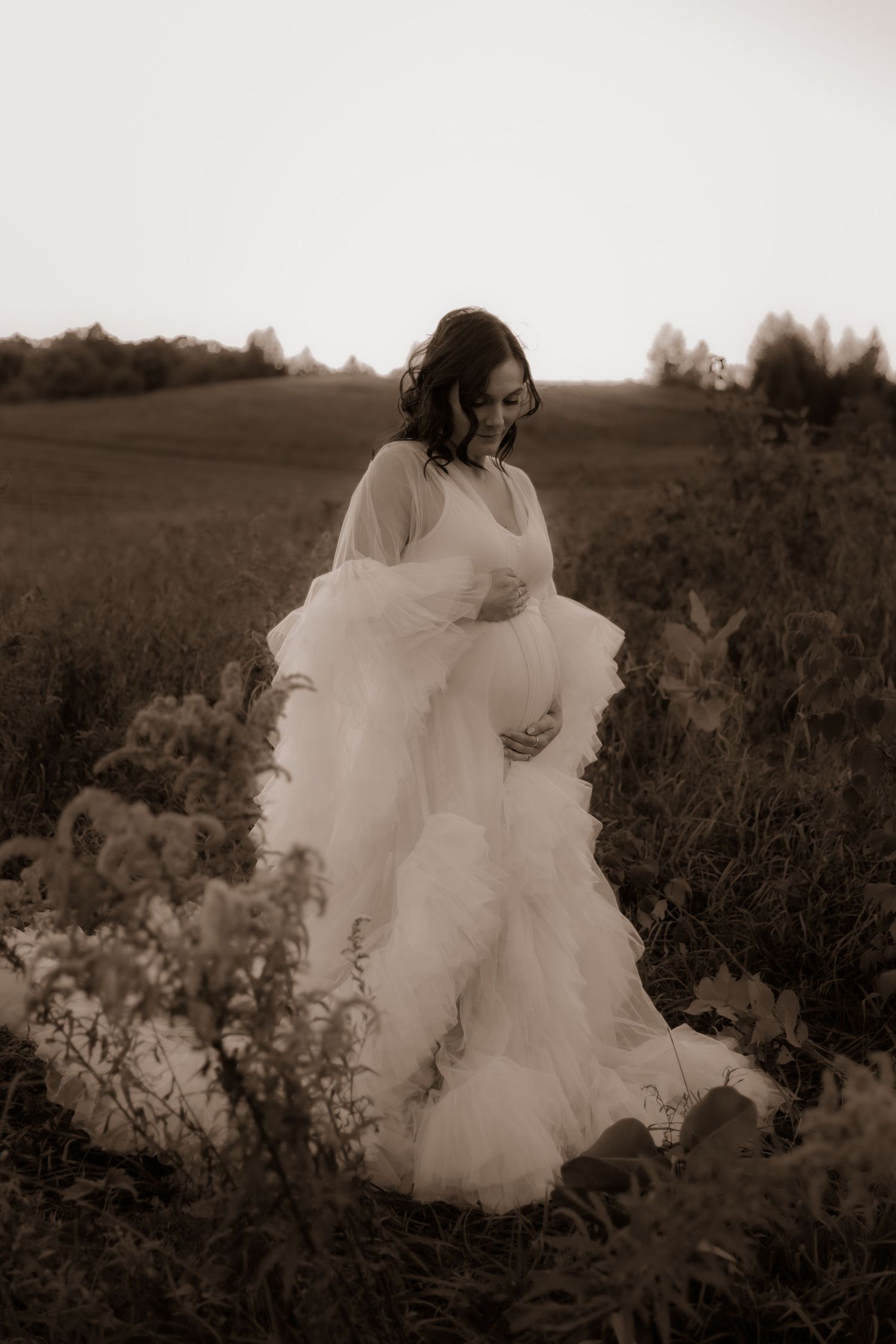 A romantic sepia-toned bridal portrait showing a flowing white wedding dress among wildflowers in a rustic field.