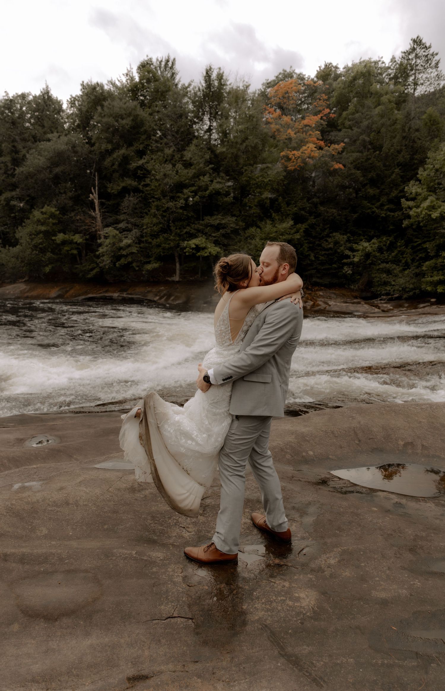 Newlyweds share an intimate moment on rocky terrain near a rushing stream surrounded by fall trees.
