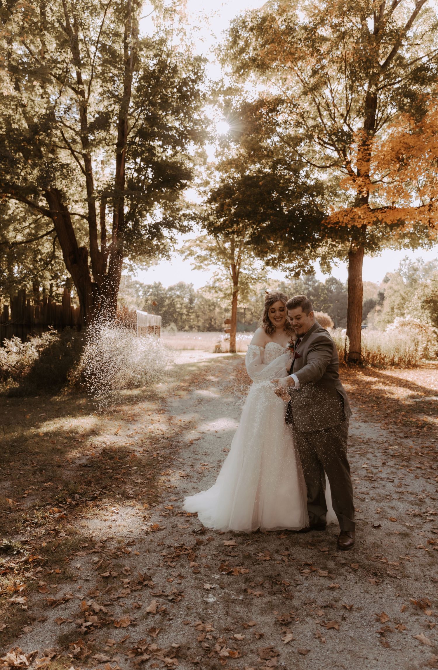 Newlyweds share romantic moment on tree-lined path in autumn sunlight.