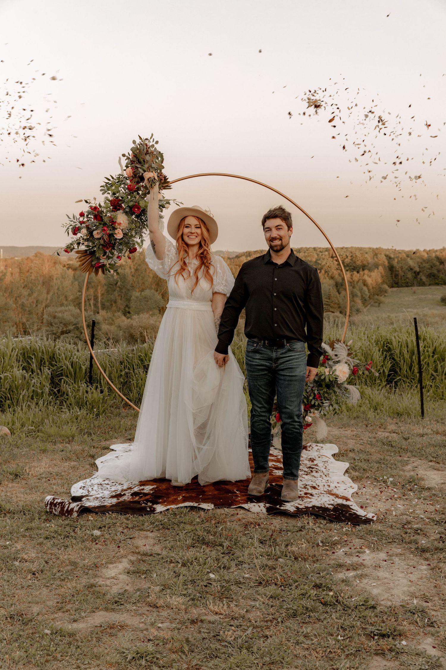 Celebratory moment under floral arch with confetti flying in golden evening light.