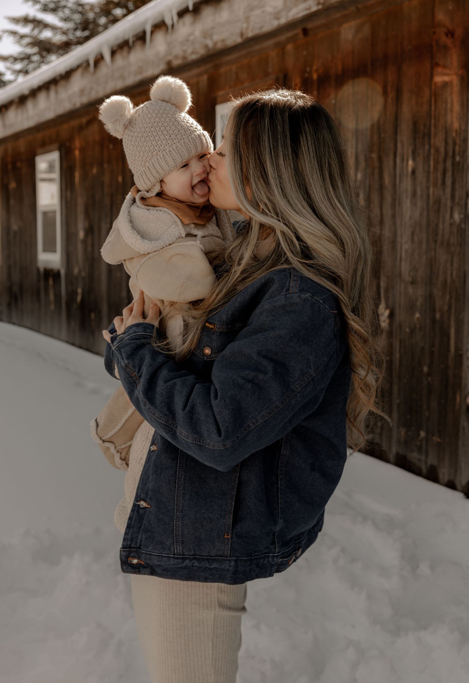 A sweet moment shared between a child in a cream beanie and parent in denim jacket against a rustic wooden cabin backdrop.