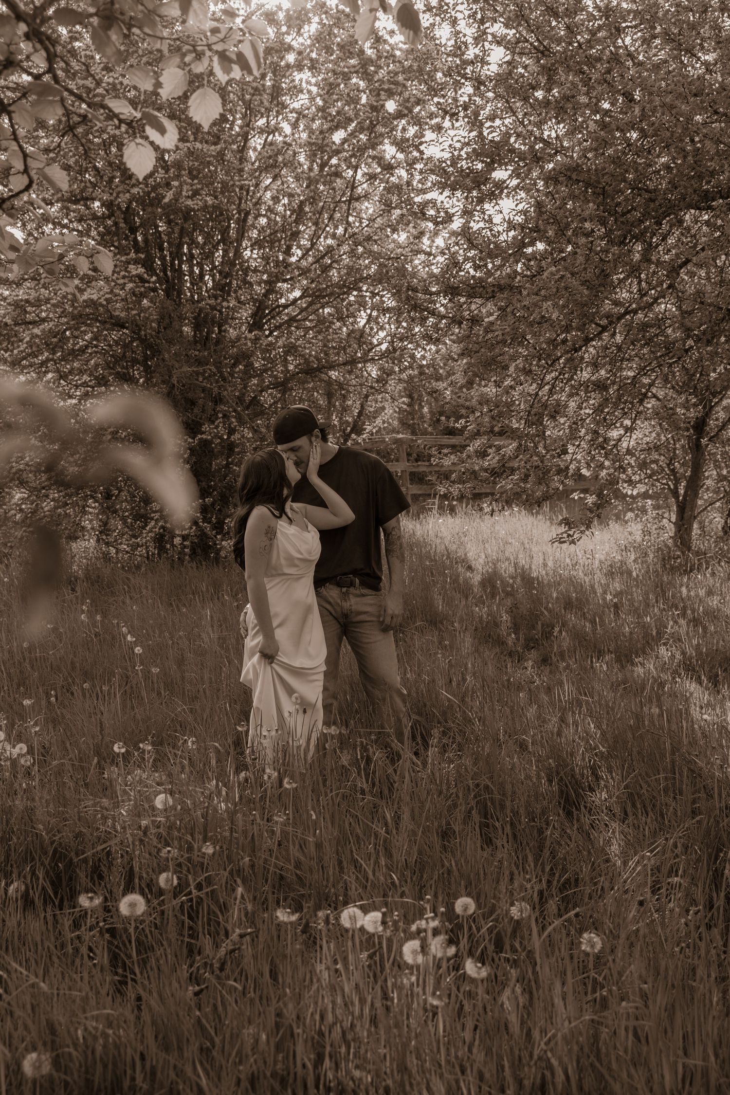 A romantic sepia-toned scene of a couple embracing in a wildflower meadow surrounded by trees on a summer day.