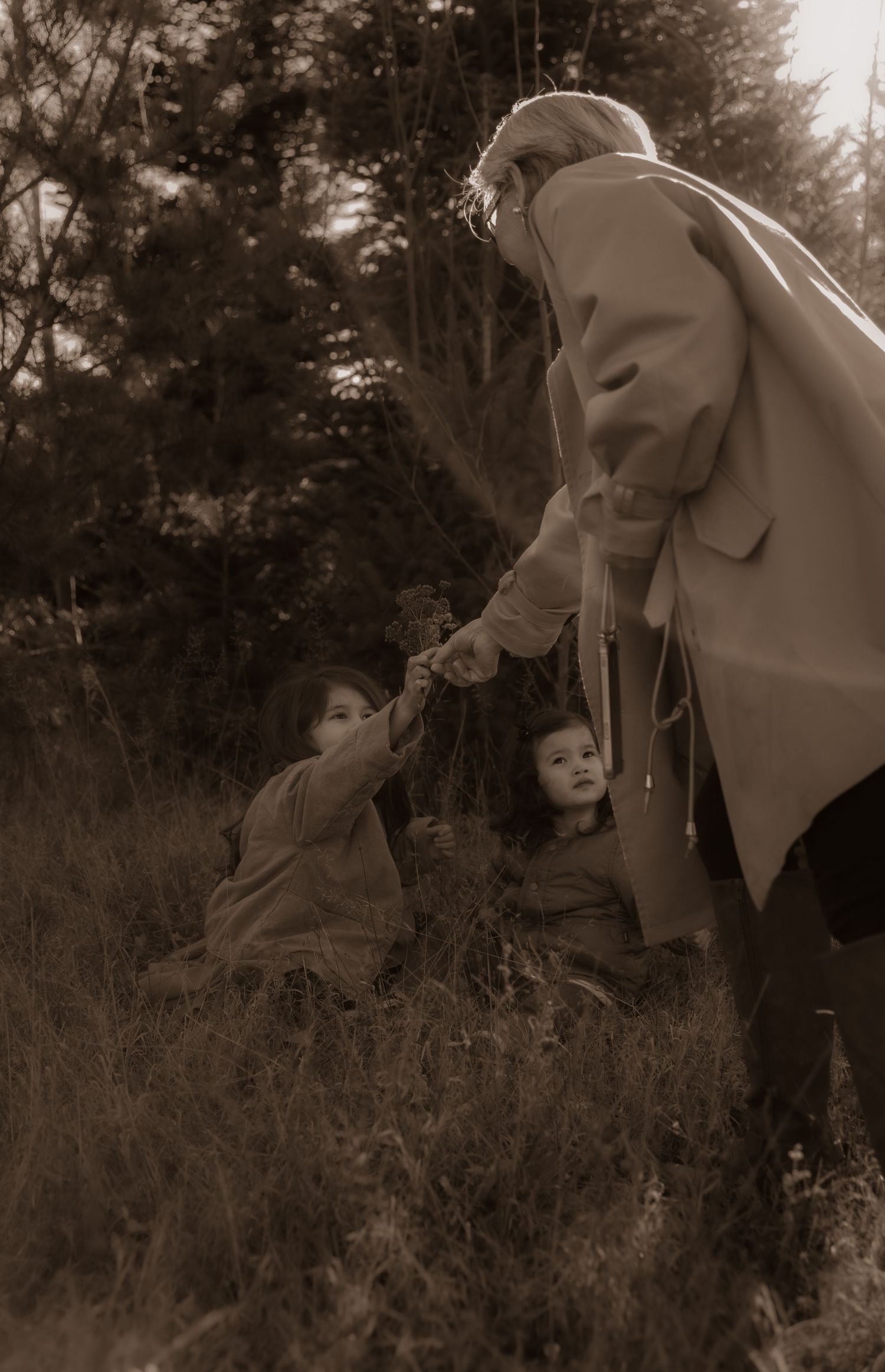 A sepia-toned photograph shows a tender moment between an older person and small children in tall grass outdoors.