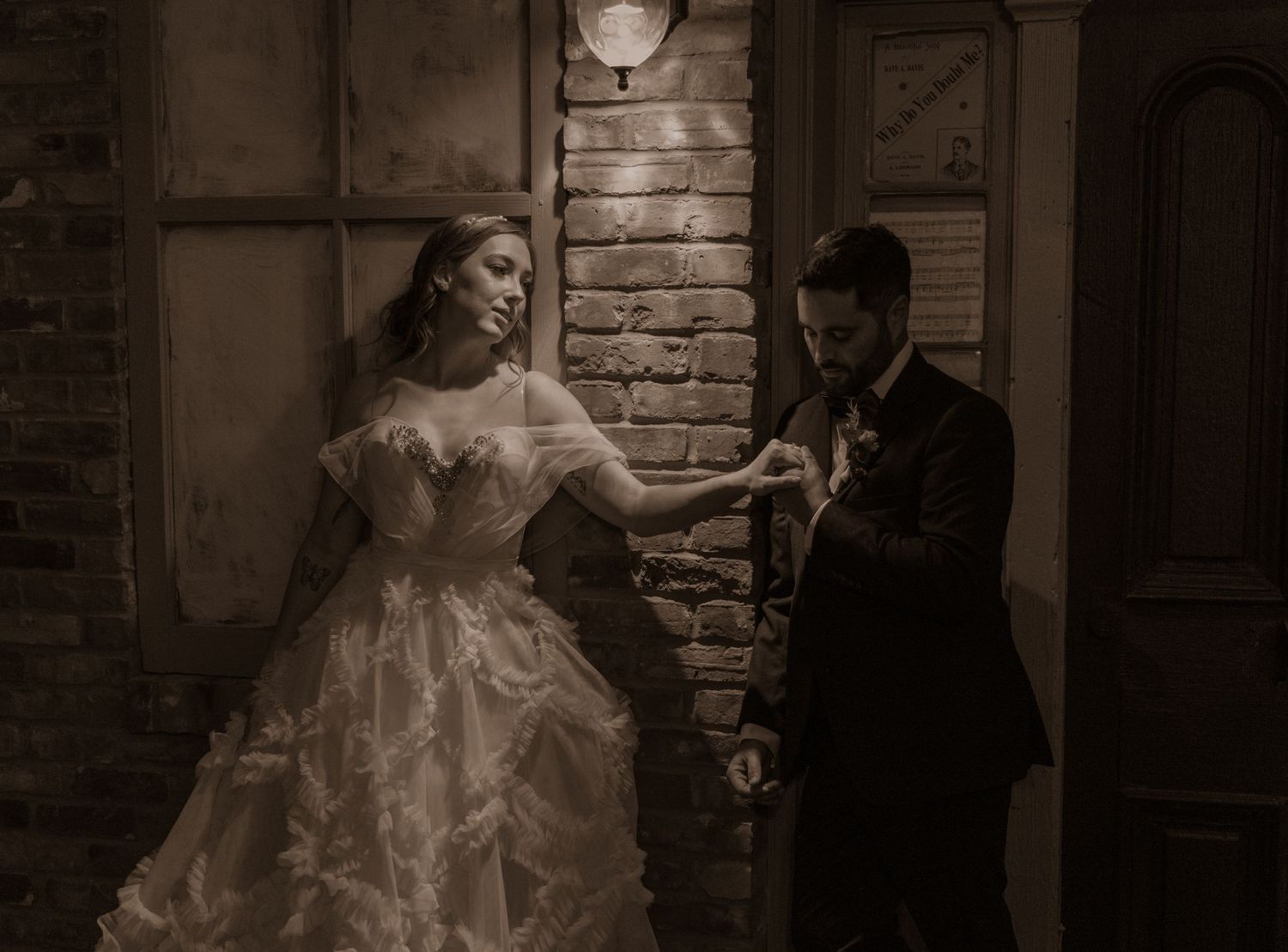 Sepia-toned wedding photo showing couple in elegant attire against brick wall with vintage lighting.