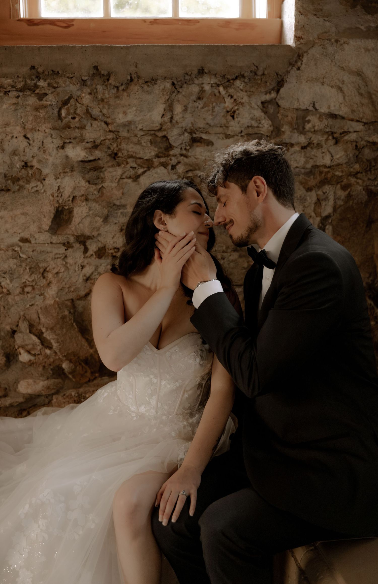Intimate wedding moment captured in warm tones against textured stone walls and natural lighting.