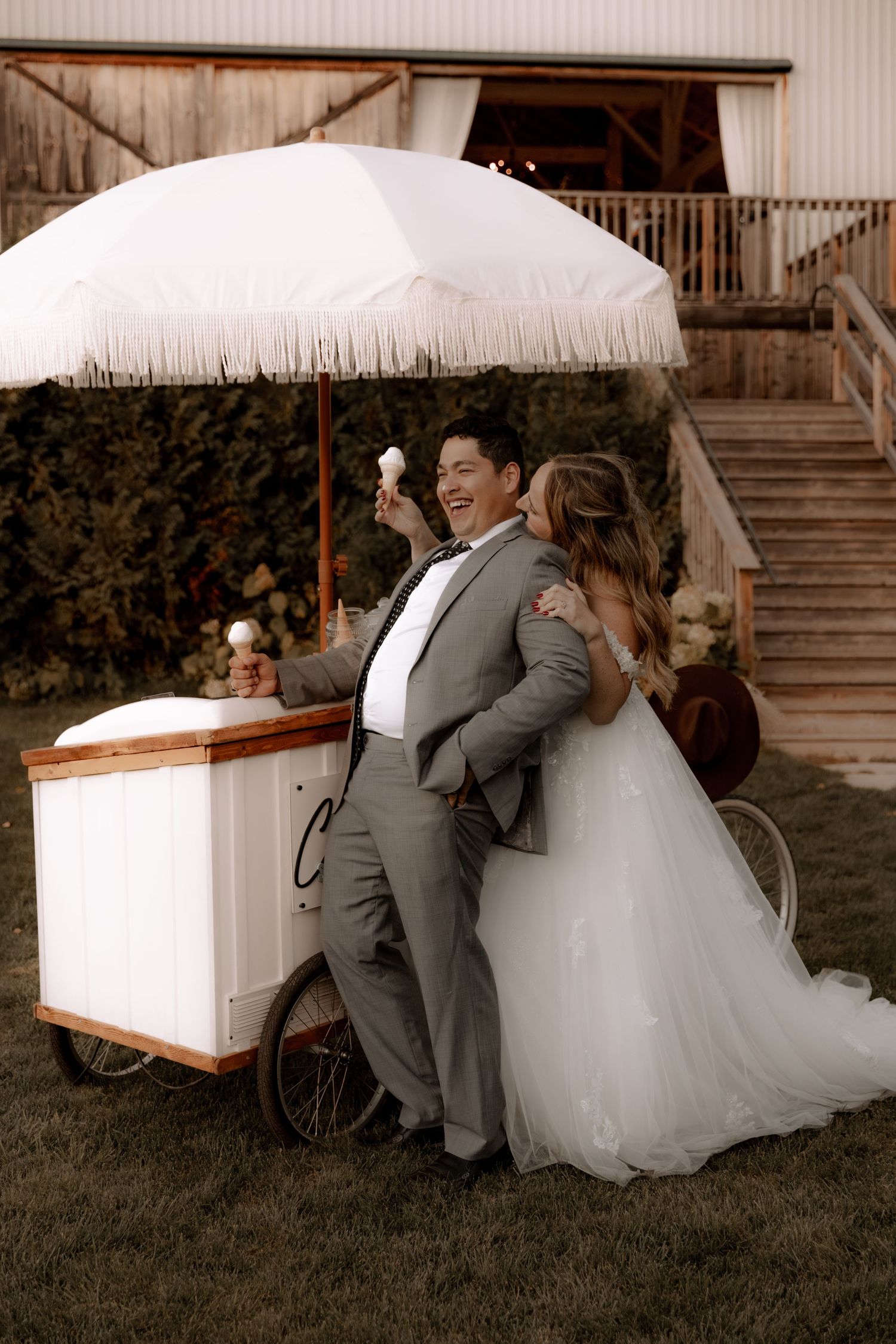 Wedding couple laughing by vintage ice cream cart under white fringe umbrella.