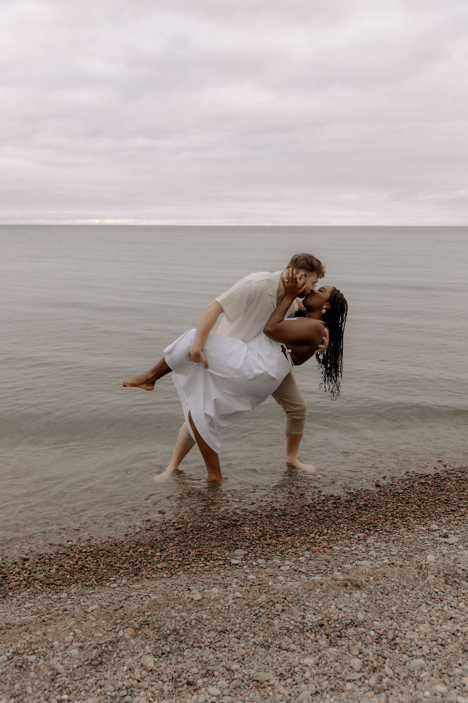 A romantic dip pose on a rocky beach with cloudy skies and water in the background.