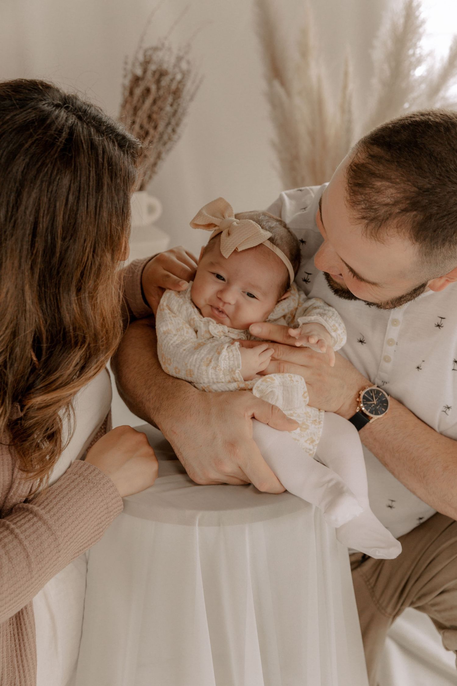 Parents tenderly hold and gaze at their sleeping baby in white lace dress while sitting in warm natural light.