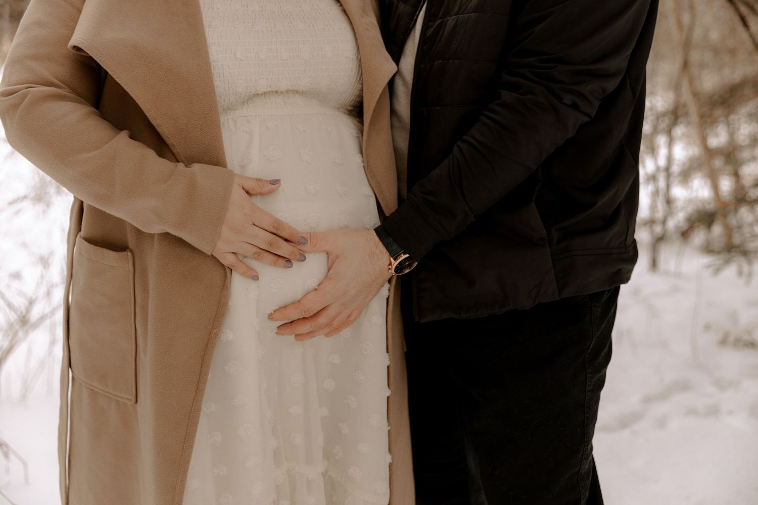 Couple shares tender moment as they cradle pregnant belly, wearing beige coat and black outfit in outdoor setting.