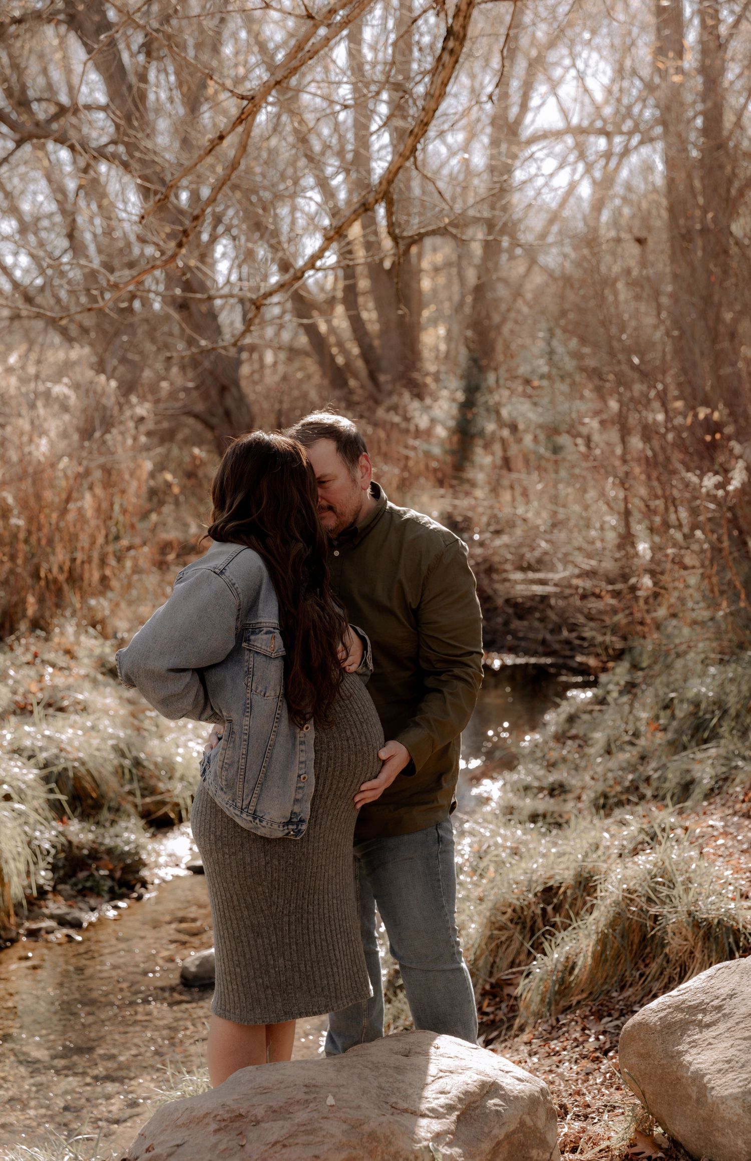 A romantic couple shares an intimate moment on a nature trail surrounded by bare autumn trees.