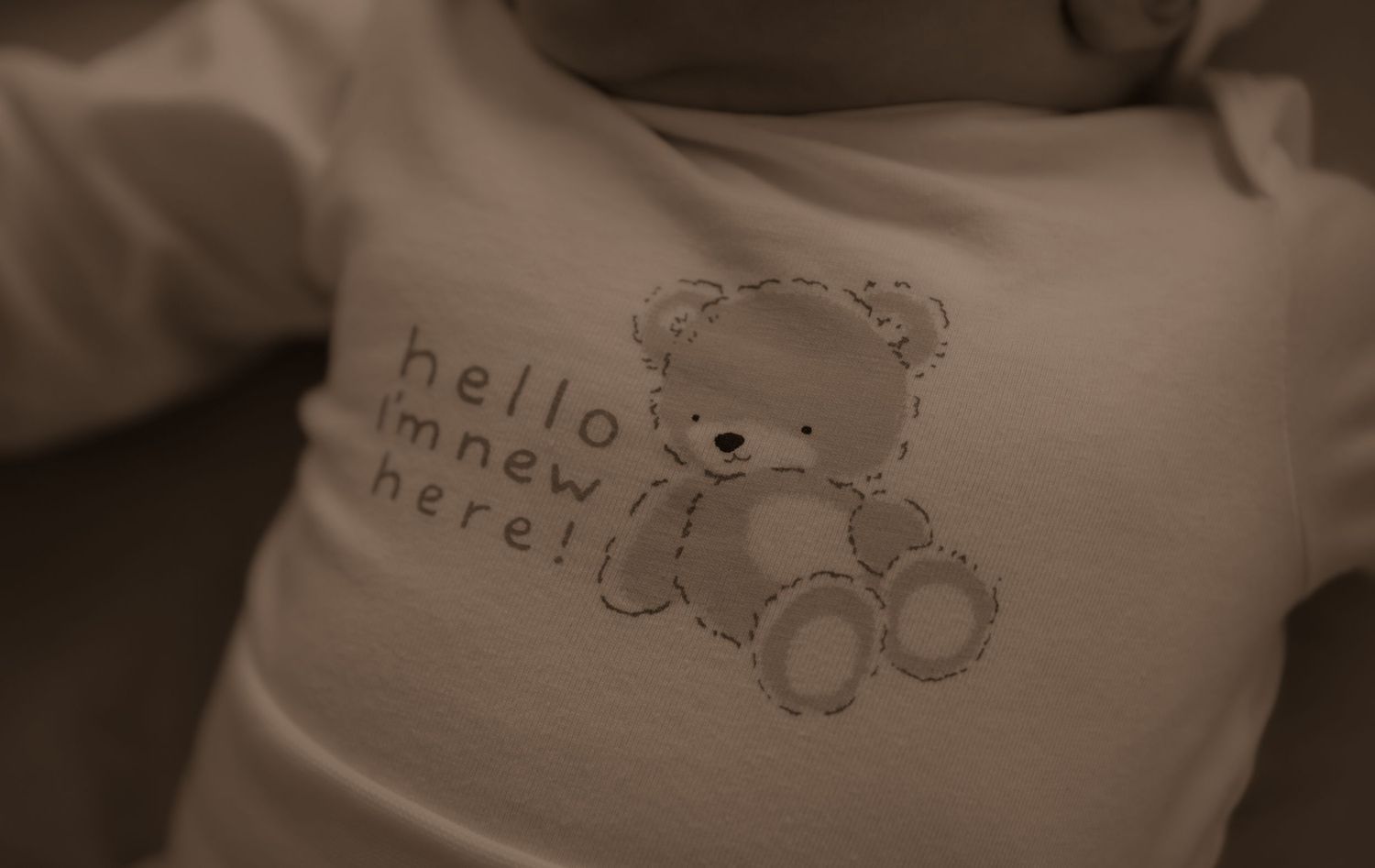 A sepia-toned close-up of a baby onesie with a cute teddy bear design and text reading 'hello inner here'.