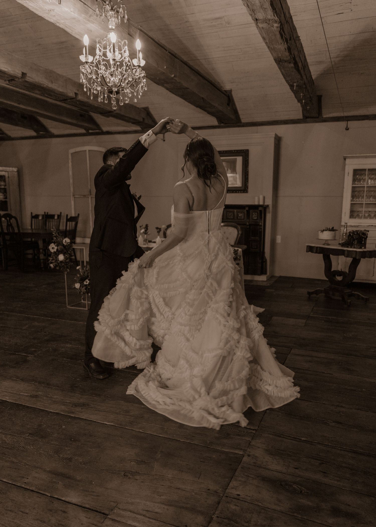 Black and white photo of newlyweds dancing under crystal chandelier in rustic venue.