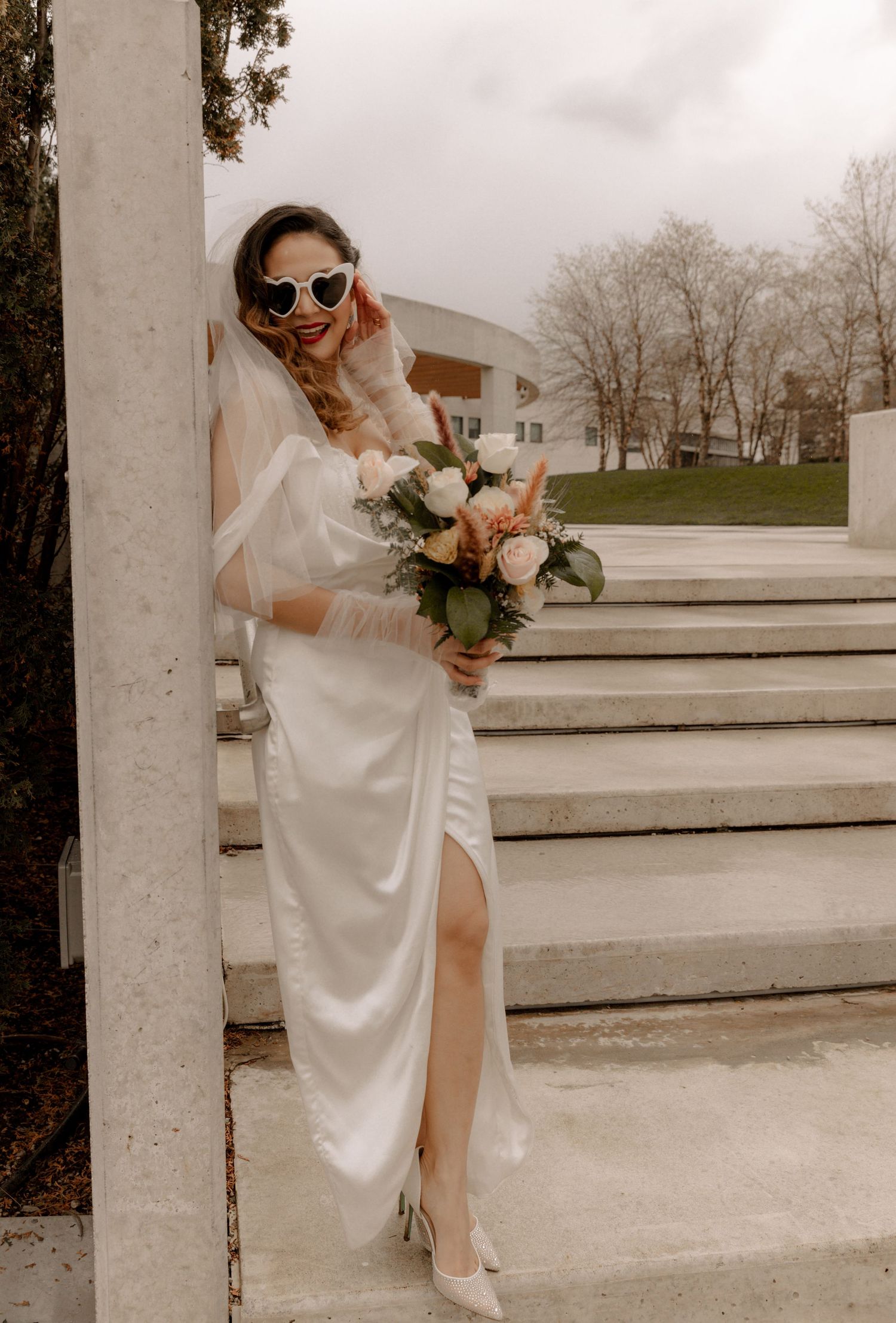 Bride in white gown and sunglasses poses with bouquet on concrete steps.