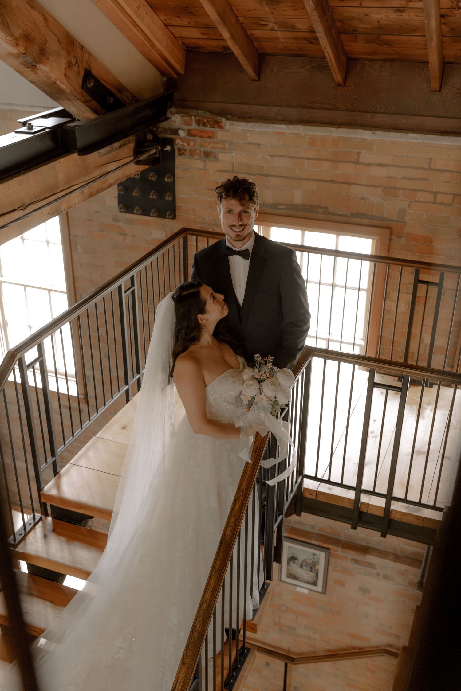 A couple embracing on a wooden staircase with metal railings in a rustic interior setting.