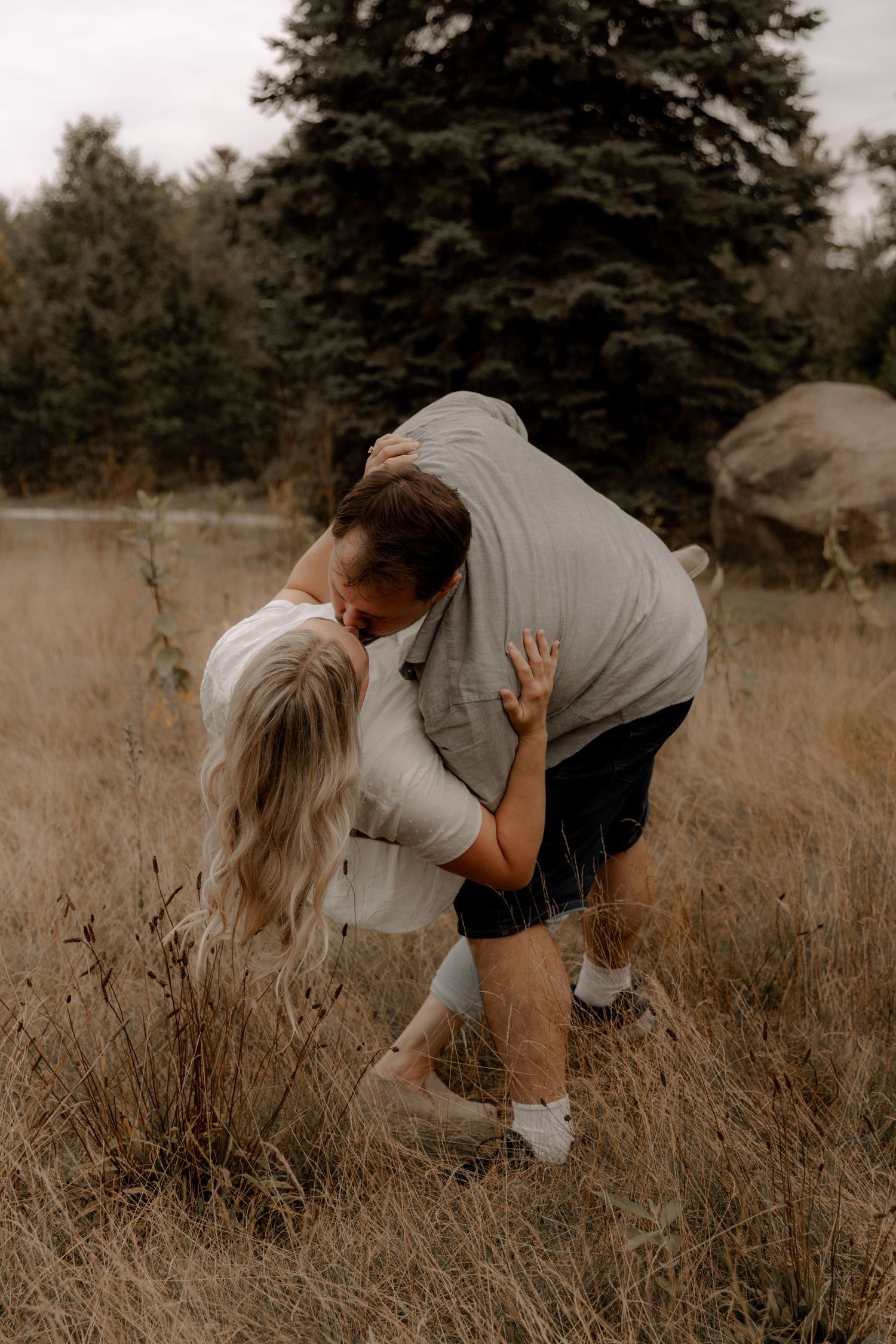 Two people share an intimate moment in a golden field beneath a large pine tree.