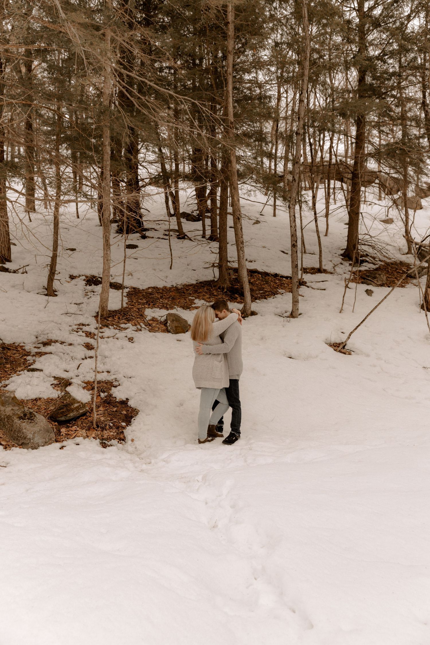 Two people embrace in a snowy winter forest landscape with bare trees and white ground cover.
