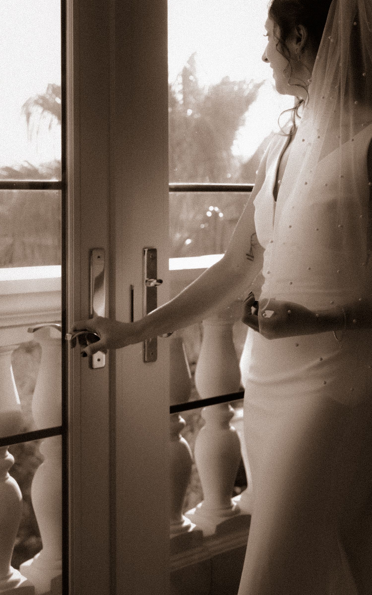 A bride in a fitted dress reaches for the handle of an elegant French door.