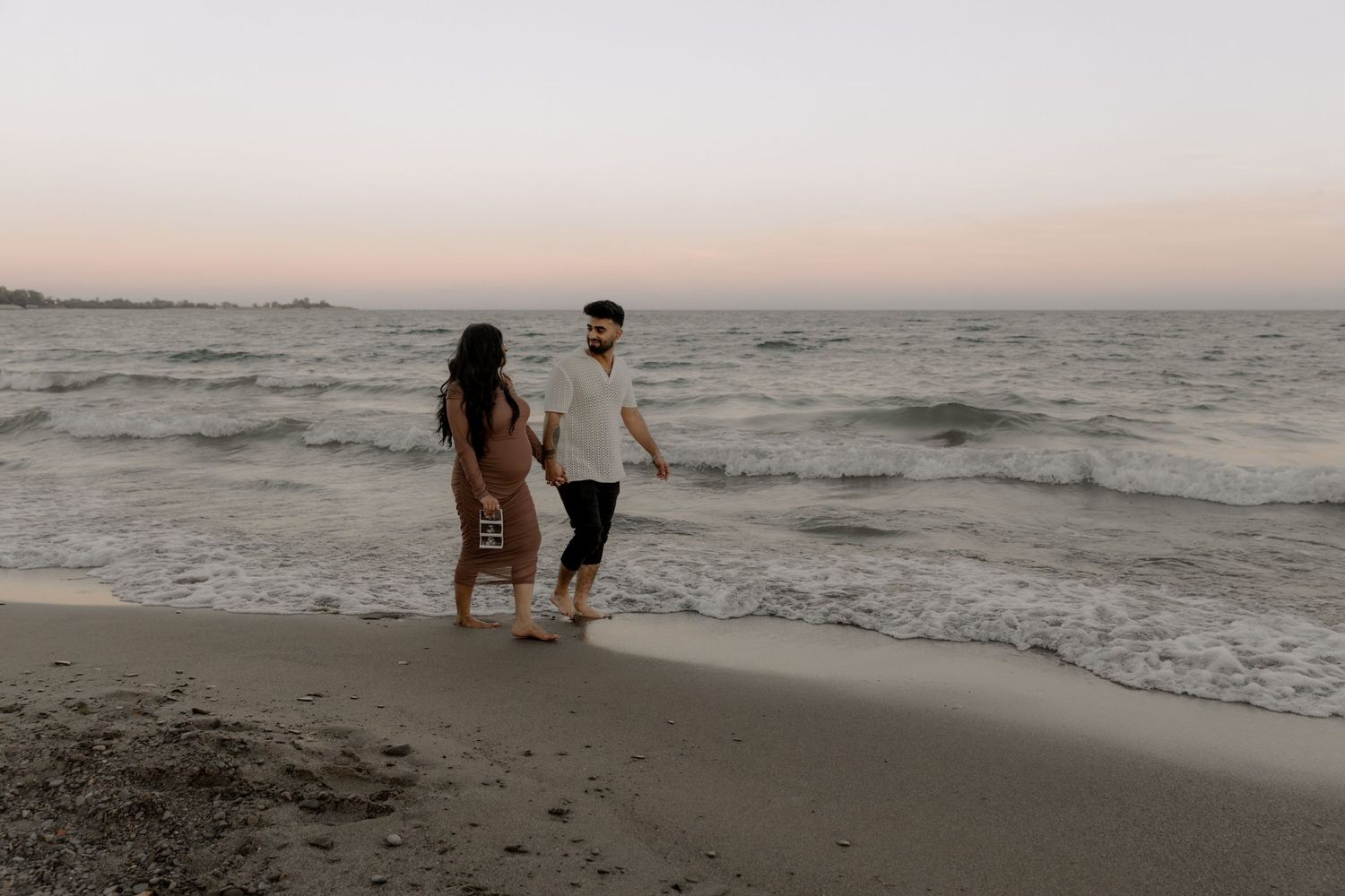 Expecting couple walking along sandy beach during sunset.