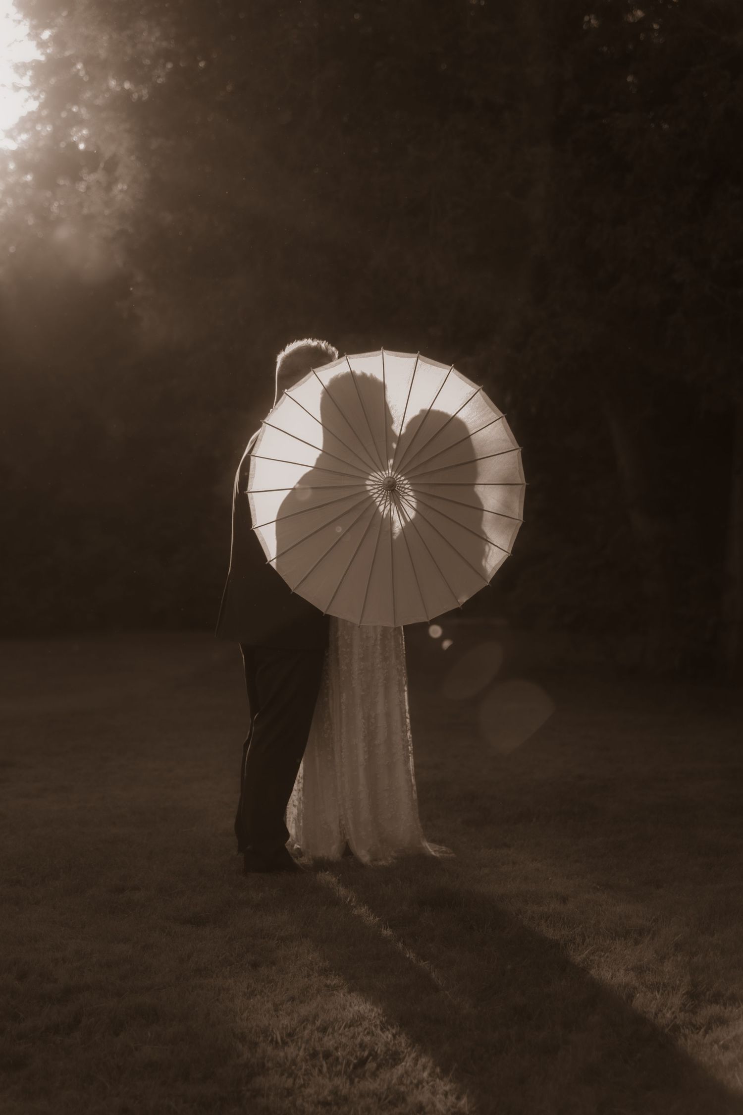 A romantic silhouette of a couple sharing a white parasol in a misty garden, captured in sepia tones.