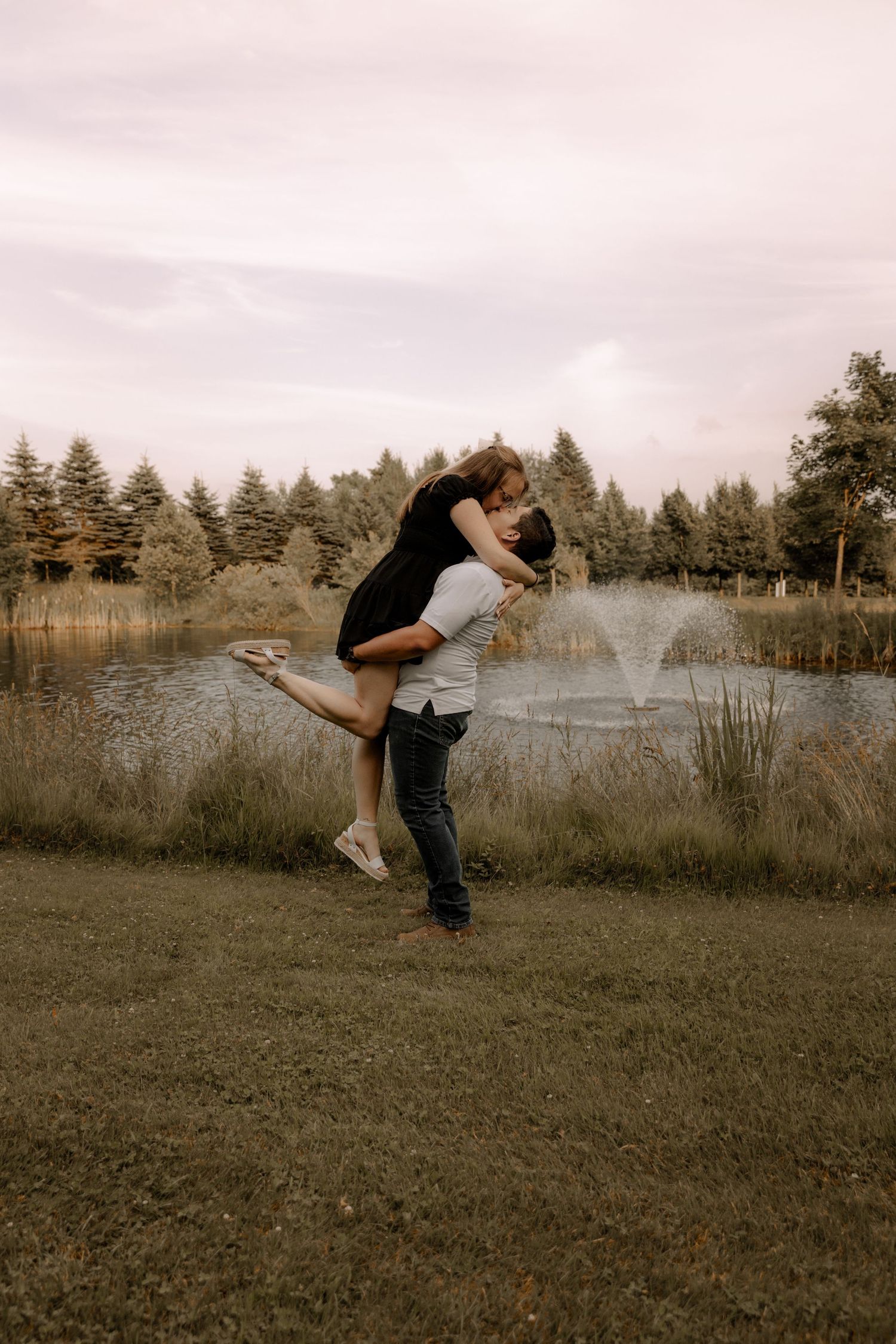 A couple shares an intimate moment beside a pond with tall pine trees framing the romantic scene.