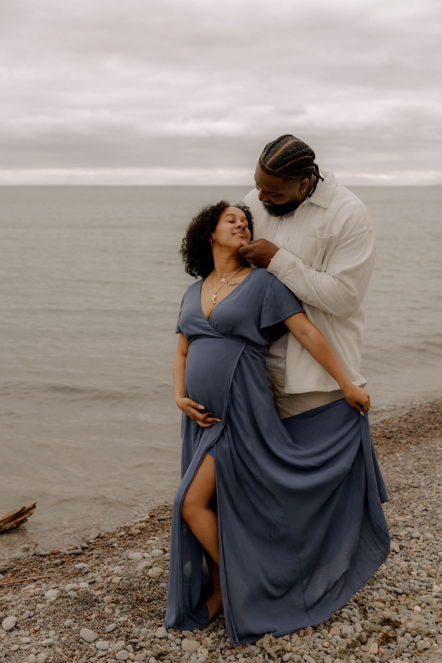 A couple shares an intimate moment on a beach, with grey skies and water in the background.