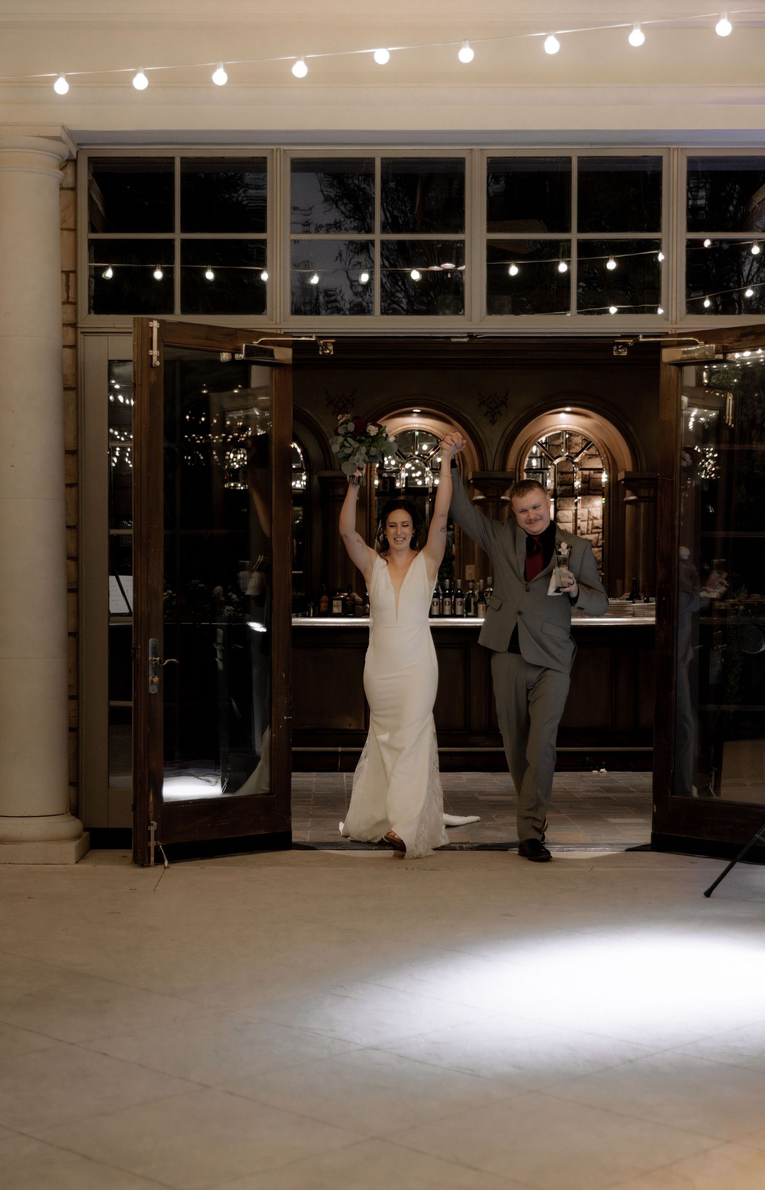 Newlyweds celebrate with raised arms under string lights at their wedding reception venue.