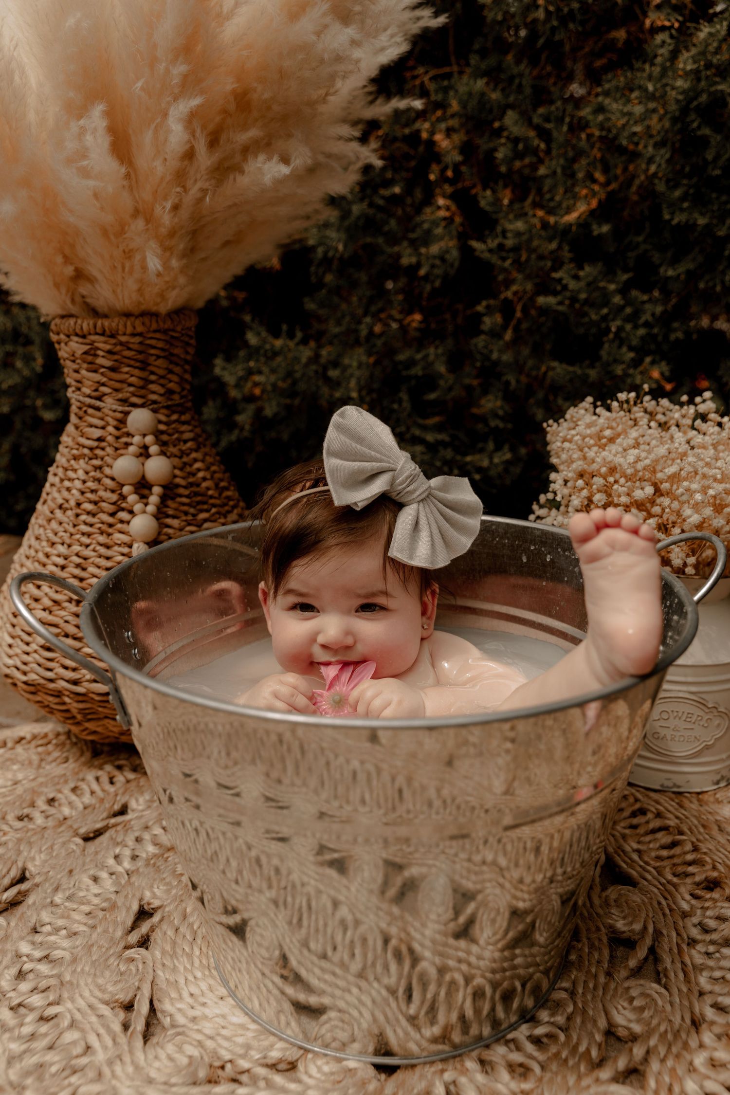 A small child sits in a decorated silver basin surrounded by boho decor including a woven vase and dried flowers.