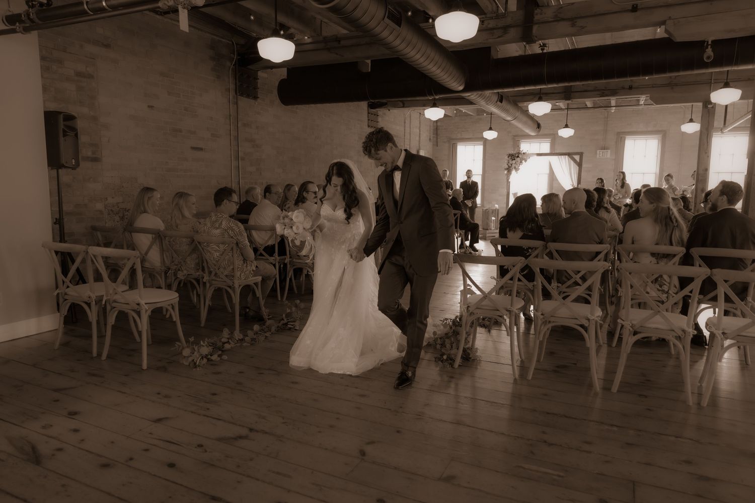 A couple walks down the aisle in a vintage-style wedding venue with industrial lighting and wooden chairs.