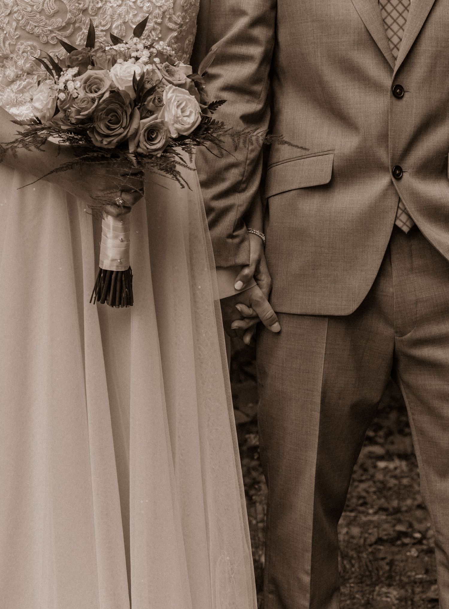 A wedding bouquet of roses held next to a beige suit jacket in a sepia-toned photograph.