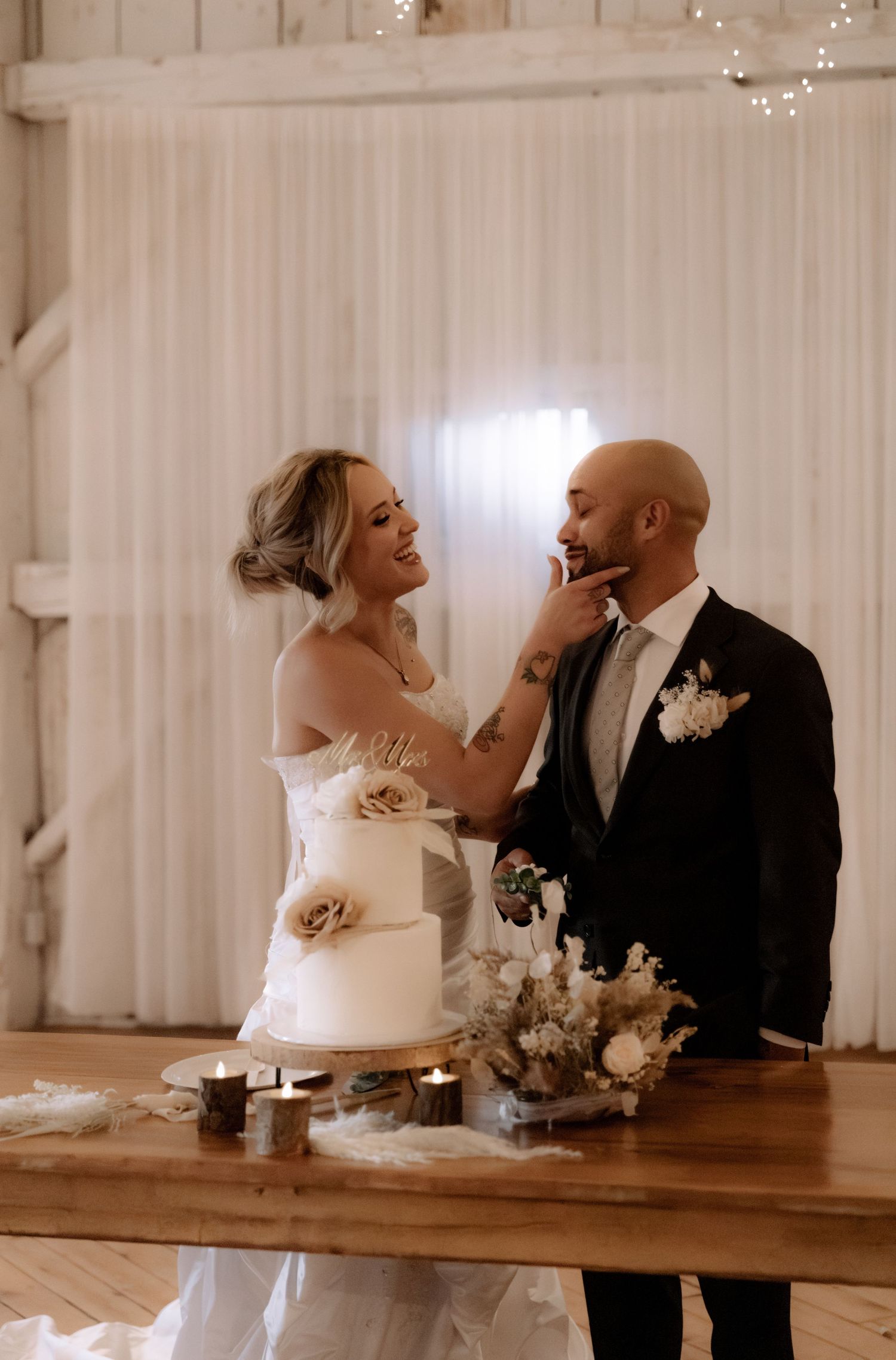 A romantic wedding cake cutting moment captured in warm lighting with a white draped backdrop and floral decorations.