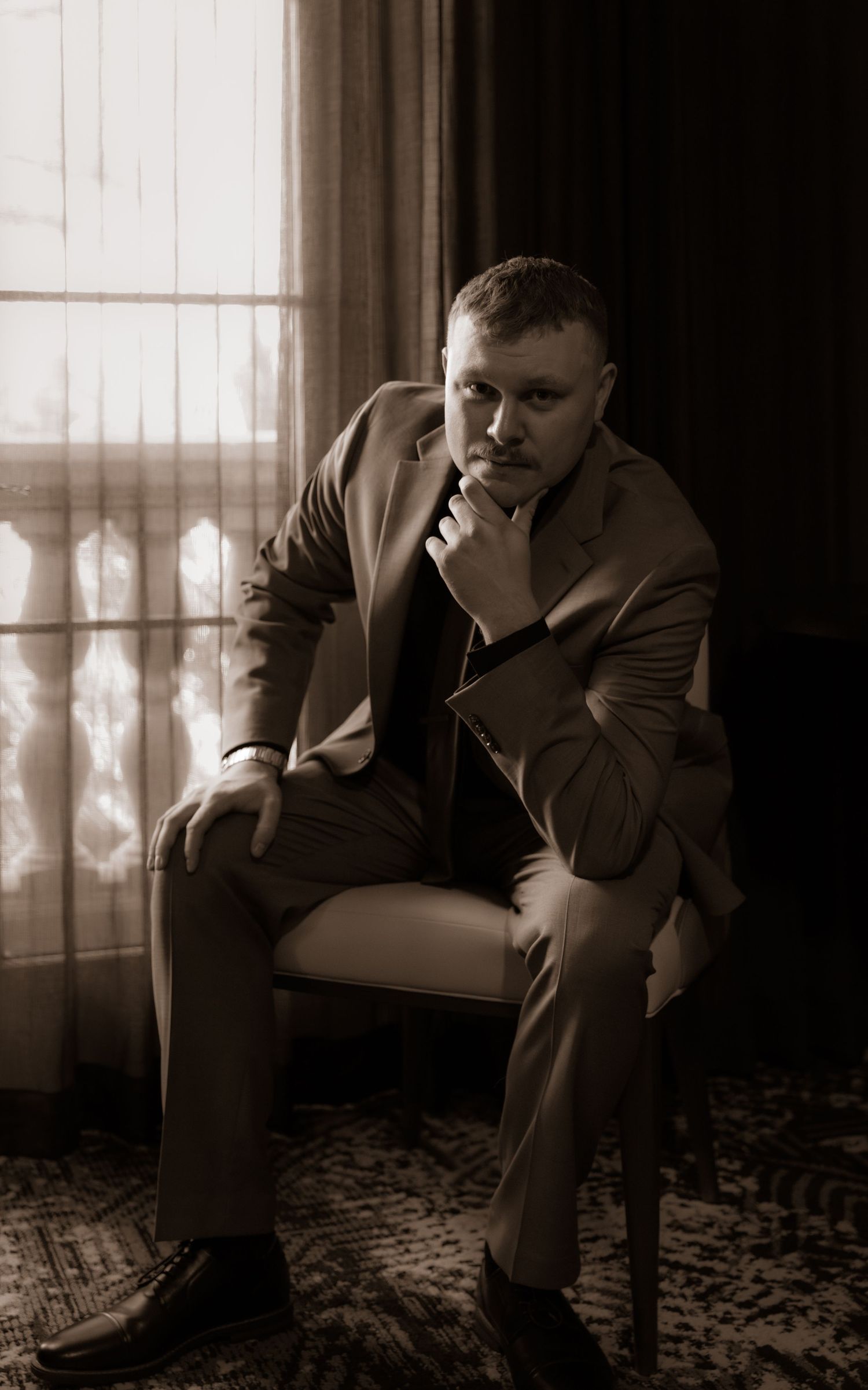 A pensive portrait in sepia tones showing someone seated in formal attire by a window.
