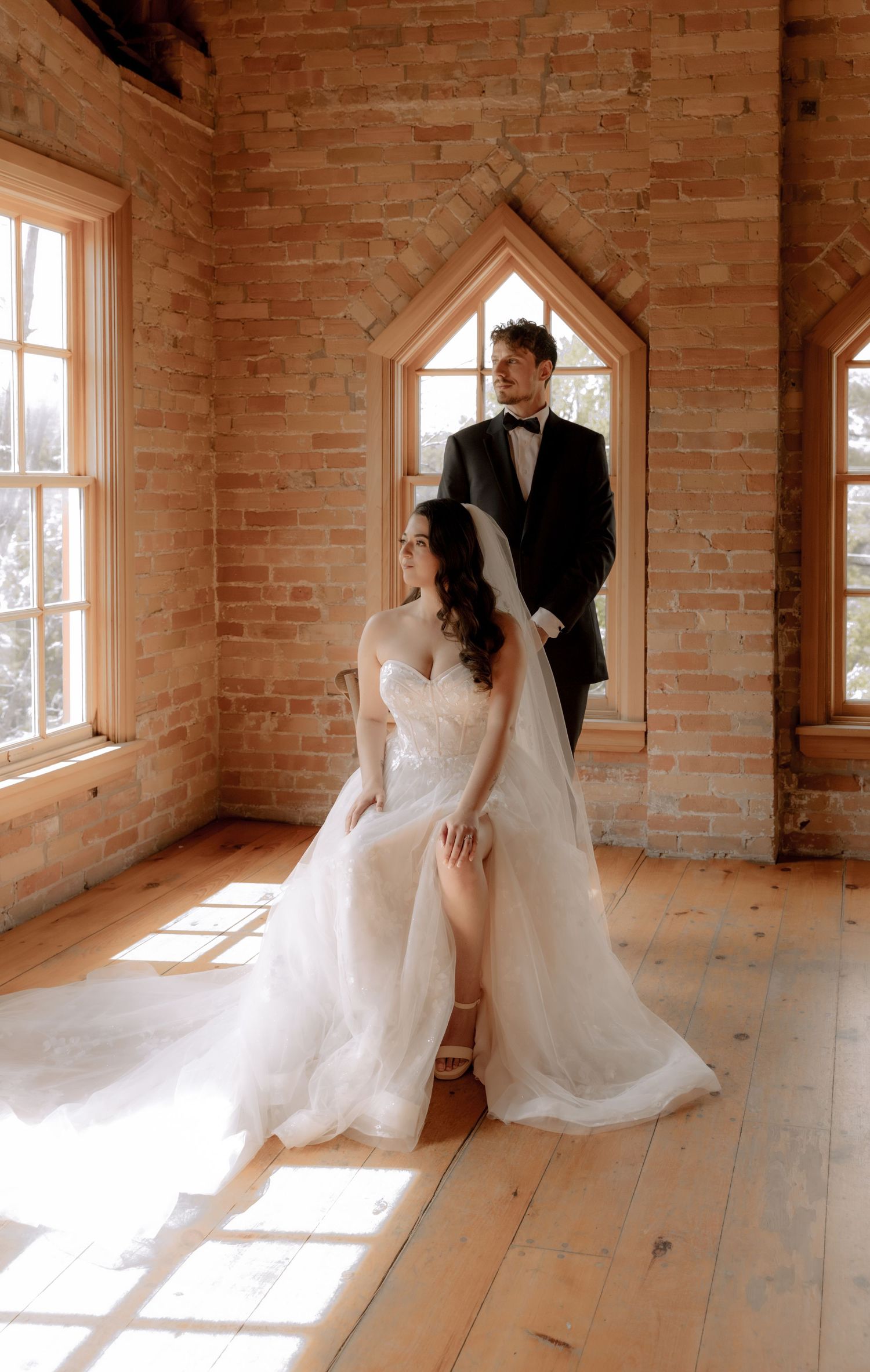 A wedding couple poses in elegant attire inside a rustic brick room with arched window alcoves.
