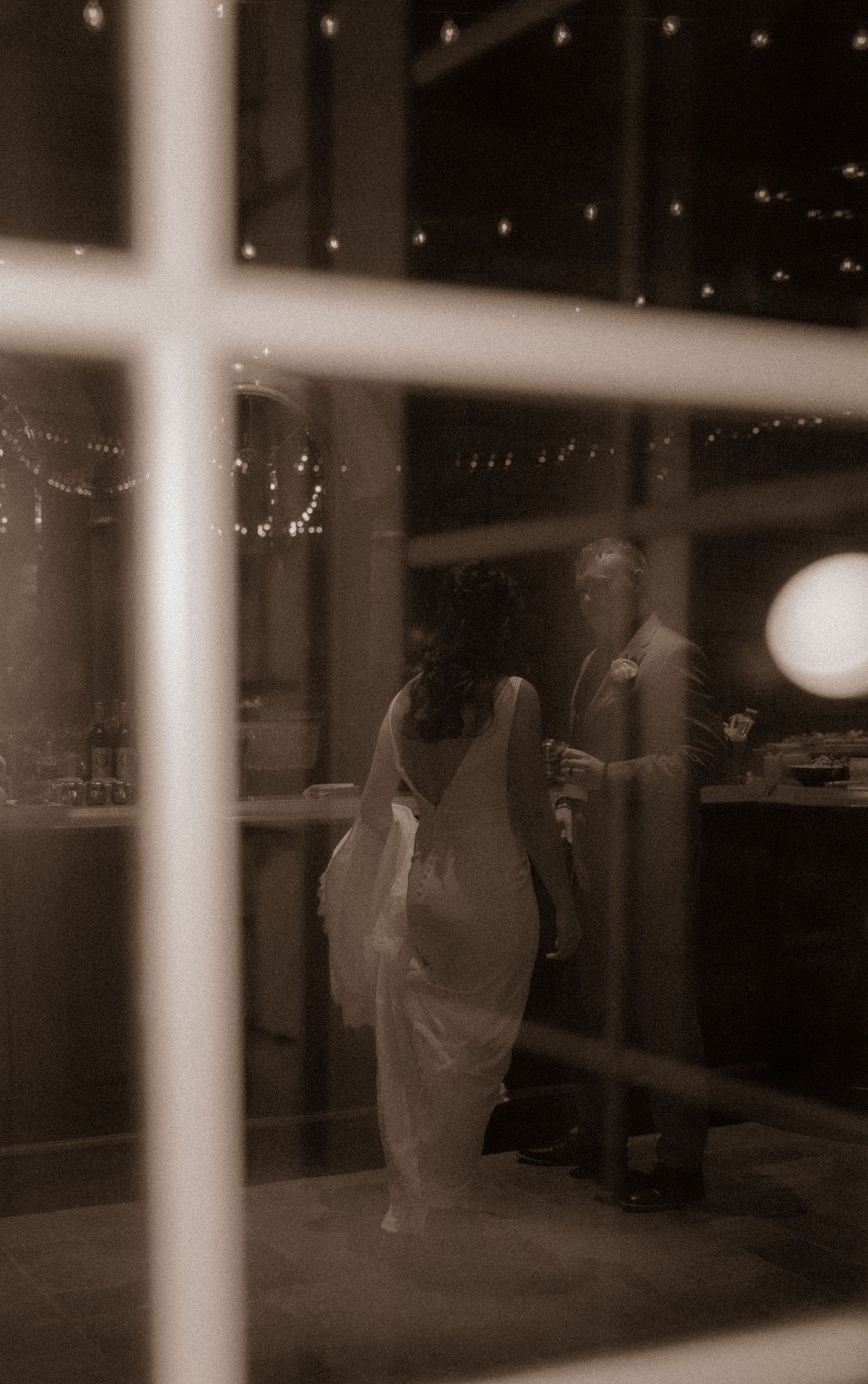 Sepia toned image of a couple dancing at an indoor reception viewed through window panes.