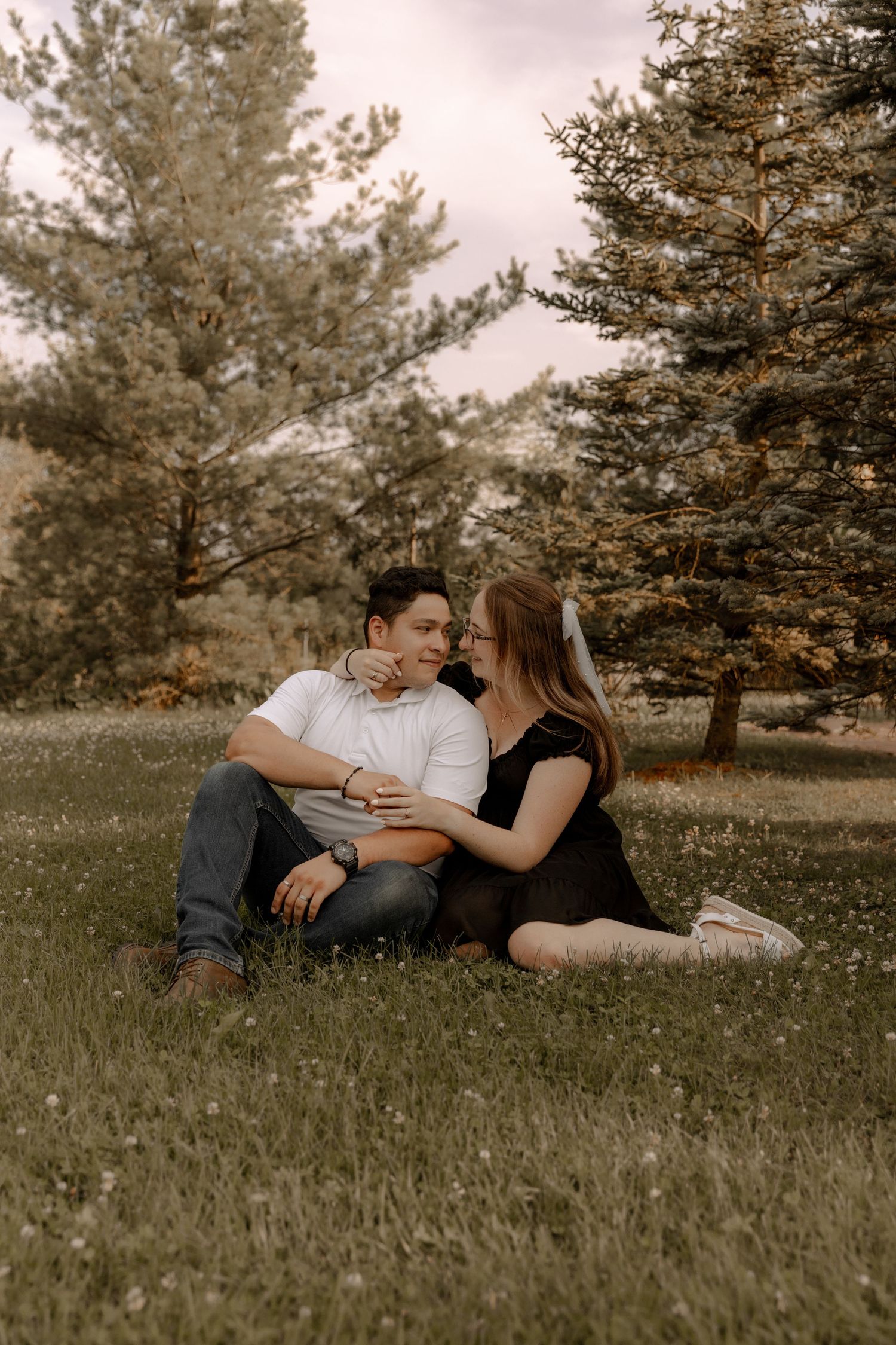 A tender moment between a couple sitting on grass with pine trees in the background.
