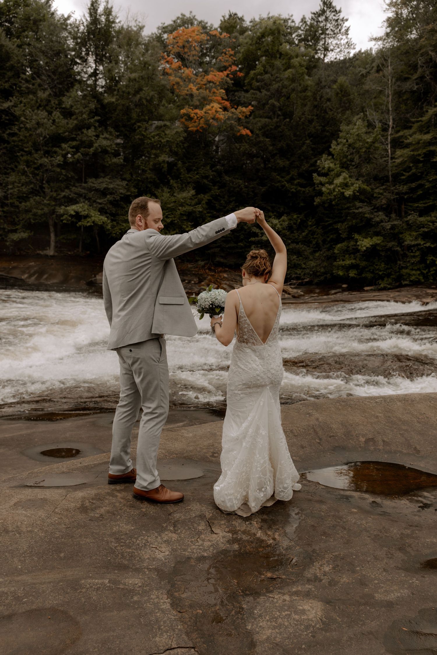 Couple in wedding attire dances on rocky riverbank with rushing water and autumn foliage in background.