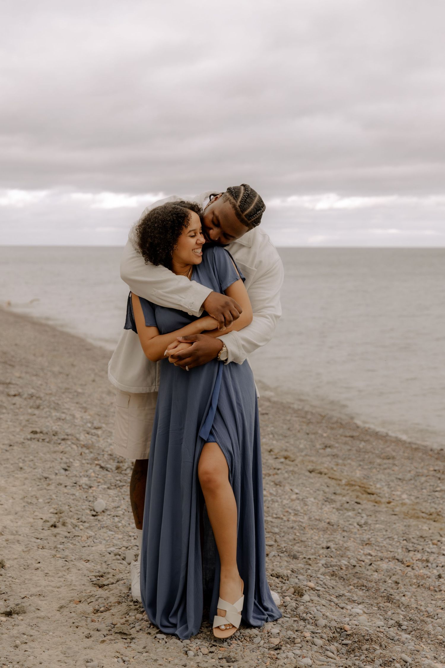 A loving couple embraces on a cloudy beach, the person wearing a flowing blue dress and the other in cream colored clothing.